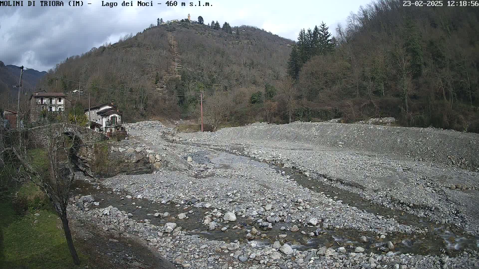 A rocky, mostly dry riverbed with a small stream flows through it, past a stone bridge and houses nestled against a hillside under a cloudy sky.