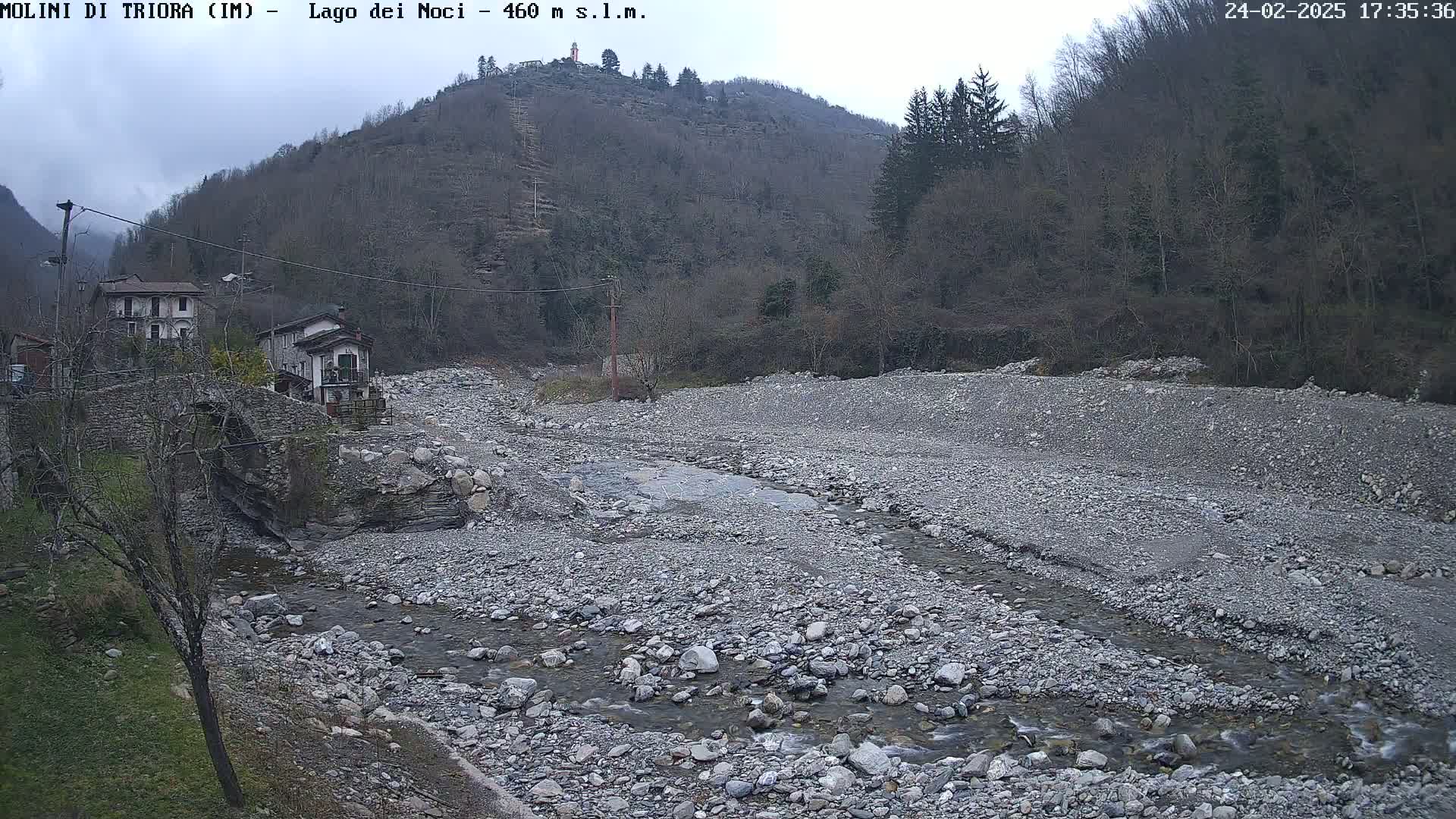 A rocky, dry riverbed with a small stream flows through it,  near buildings nestled at the base of a hill covered in leafless trees under an overcast sky.