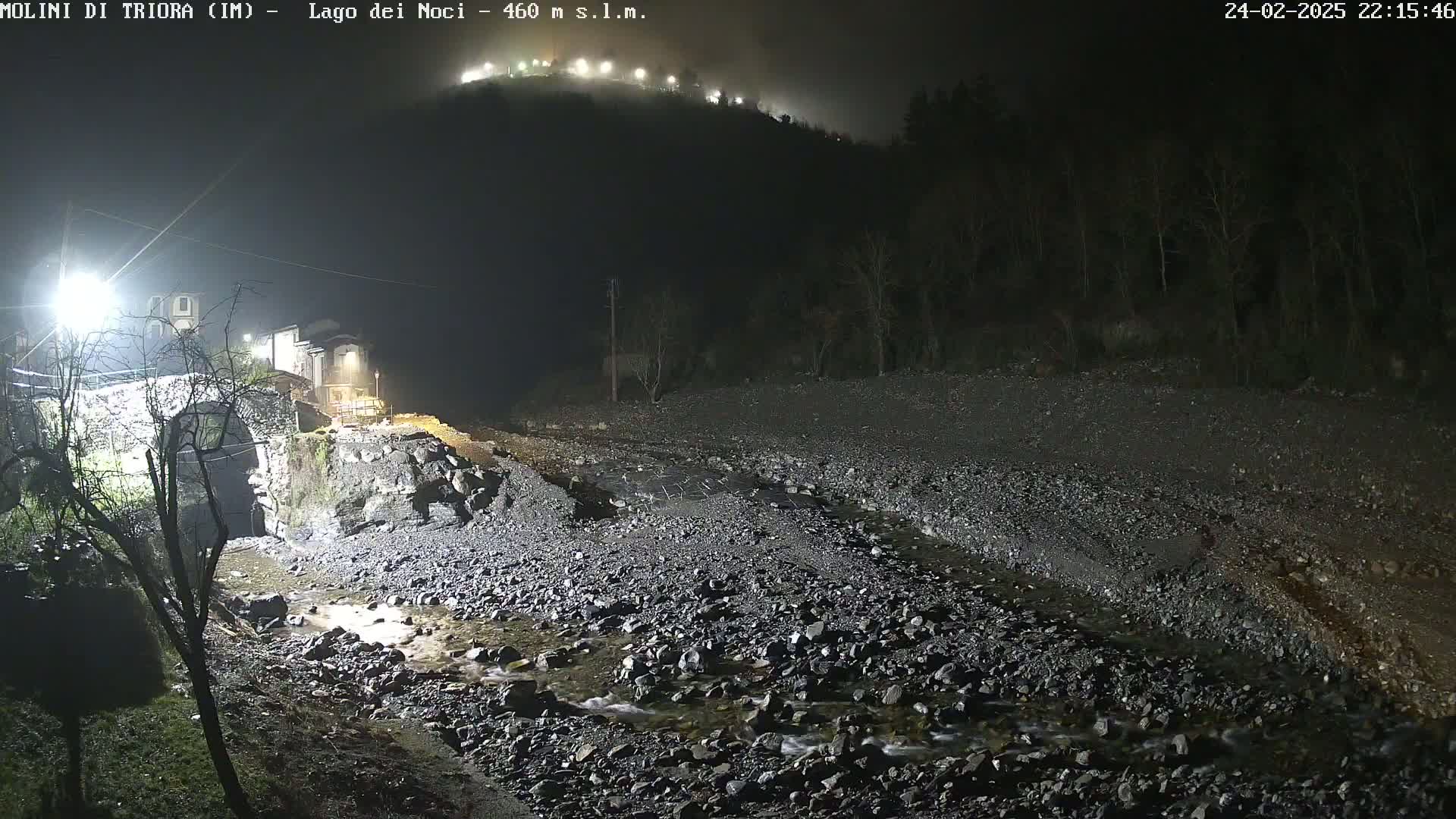 A nighttime view of a rocky streambed with a small stream flowing through it, illuminated by artificial lights, near a stone bridge and buildings under a slightly foggy sky.