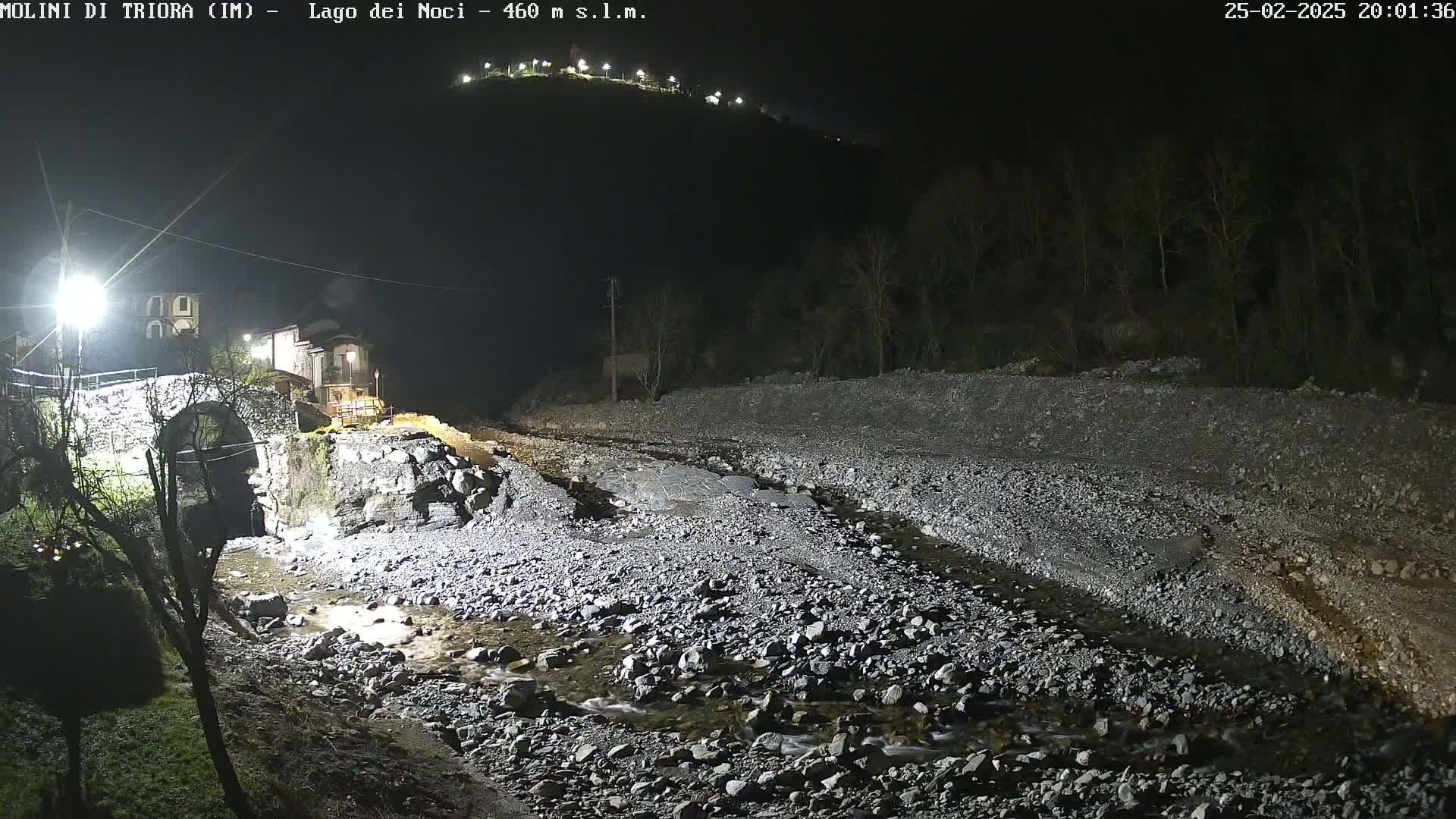 A nighttime view of a rocky, shallow riverbed under artificial light, with a stone bridge and buildings visible in the distance against a dark hillside.