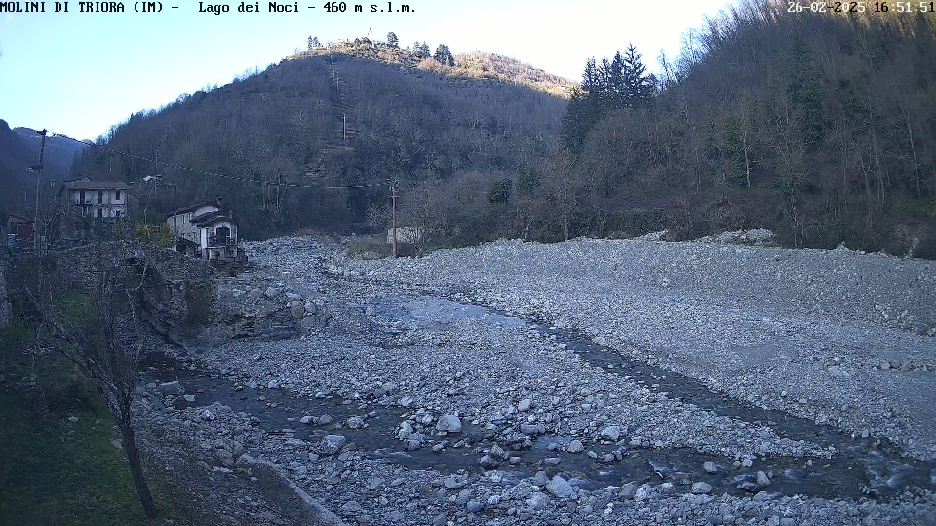 A rocky, dry riverbed with a small stream meanders through it, past a village nestled at the foot of a hill under a clear sky.