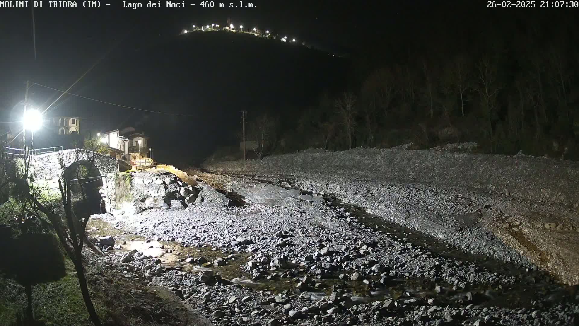 A nighttime view of a stone bridge over a shallow, rocky stream, with buildings visible in the background under artificial light.