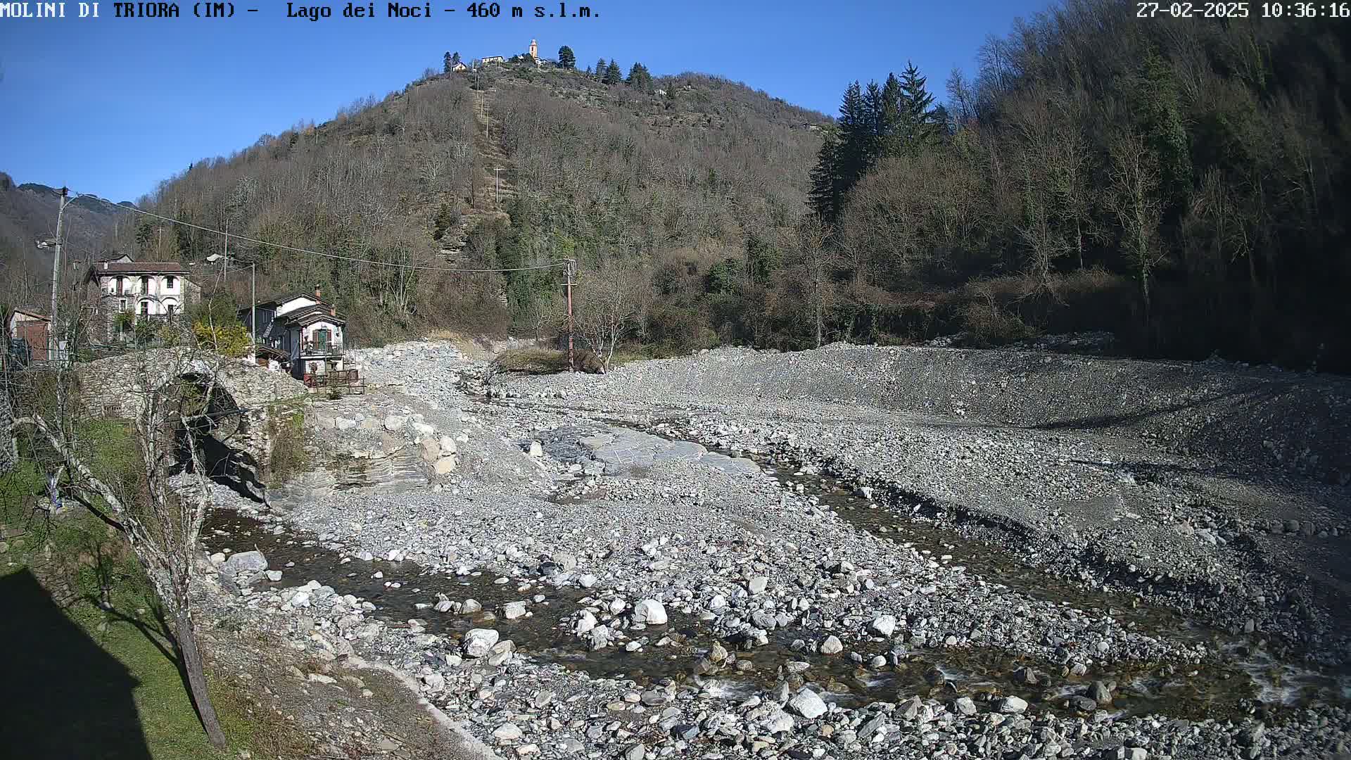 A mostly dry rocky riverbed with a small stream flows past a stone bridge and houses nestled against a hillside under a clear blue sky.