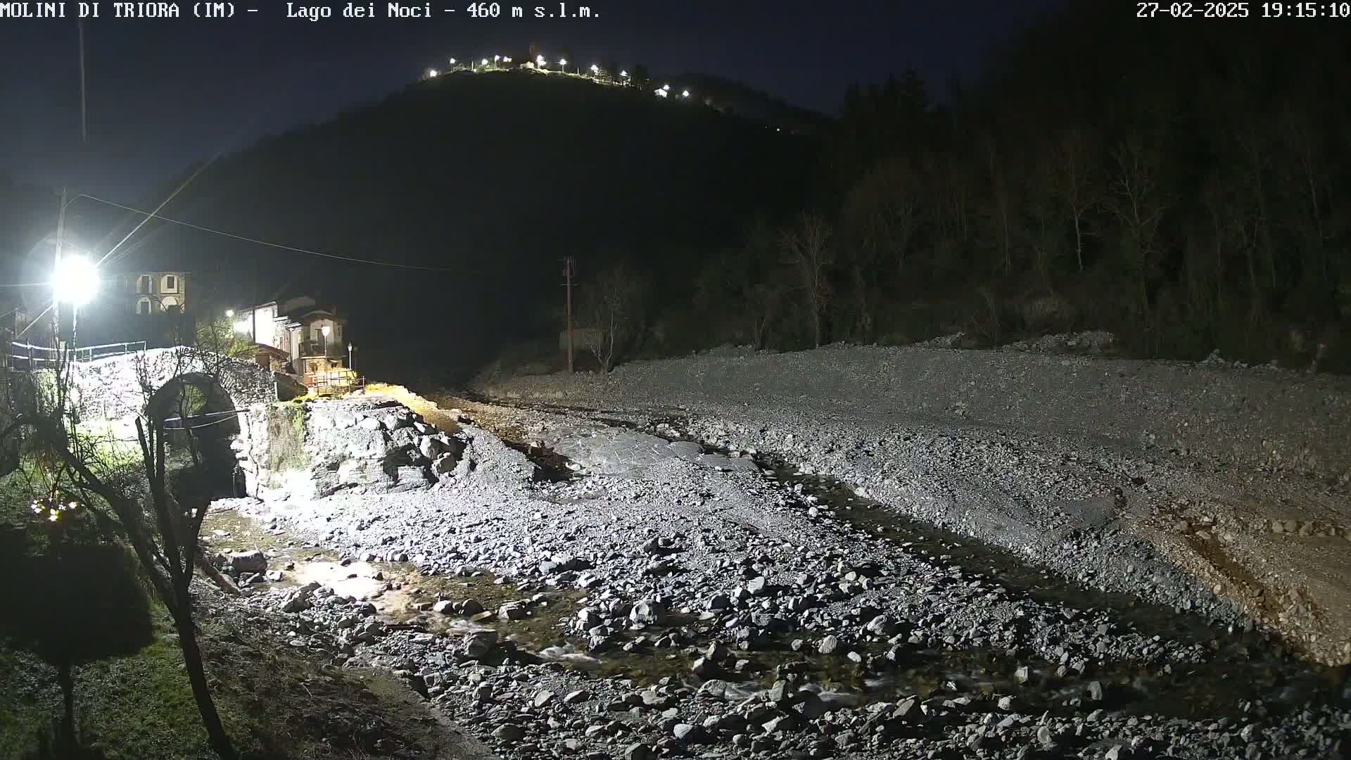 A nighttime view of a stone bridge and a rocky streambed under a dark sky, illuminated by artificial lights.