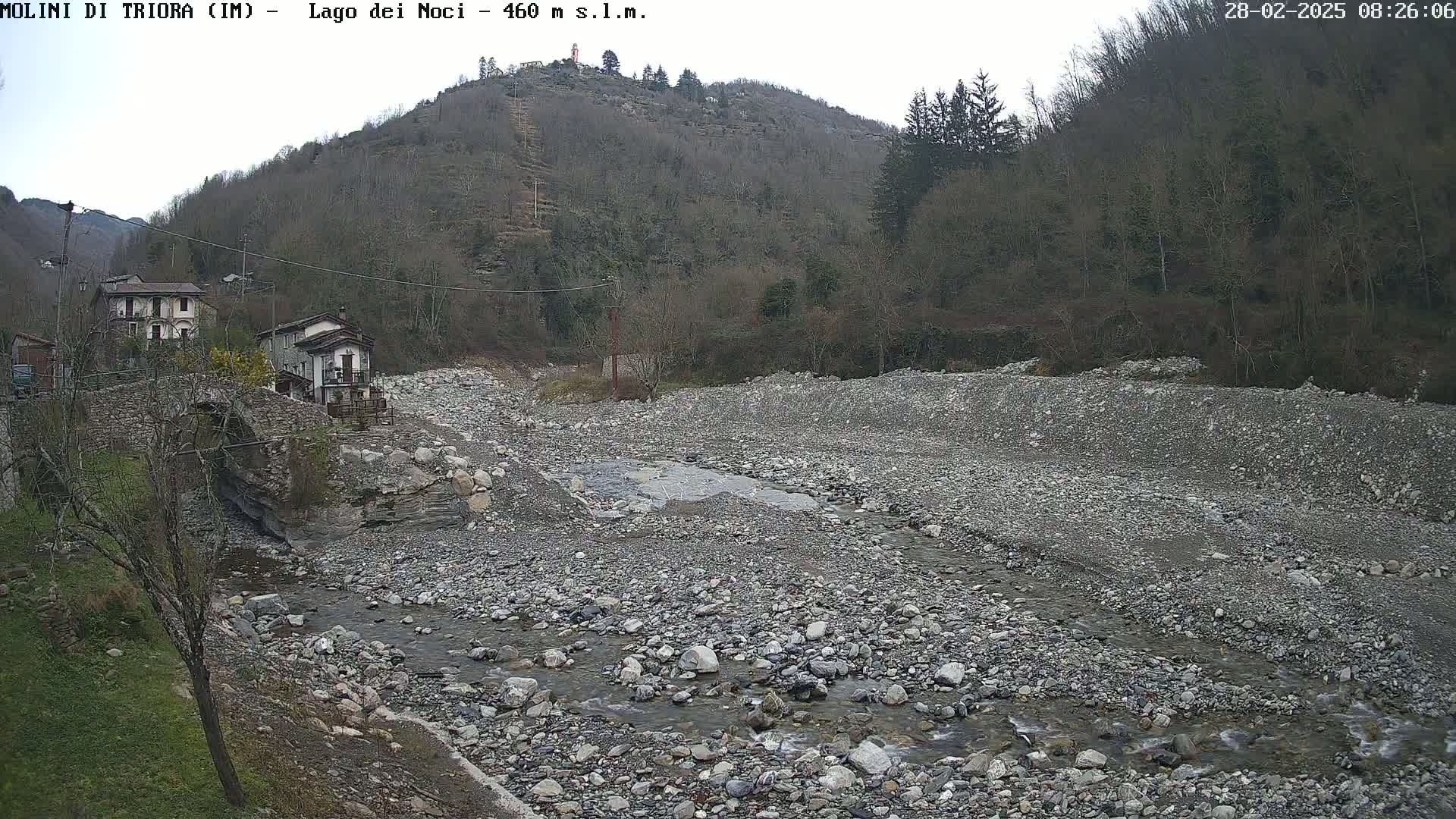A rocky, dry riverbed with a small stream flows through it, situated below a village nestled against a hillside covered in leafless trees under an overcast sky.