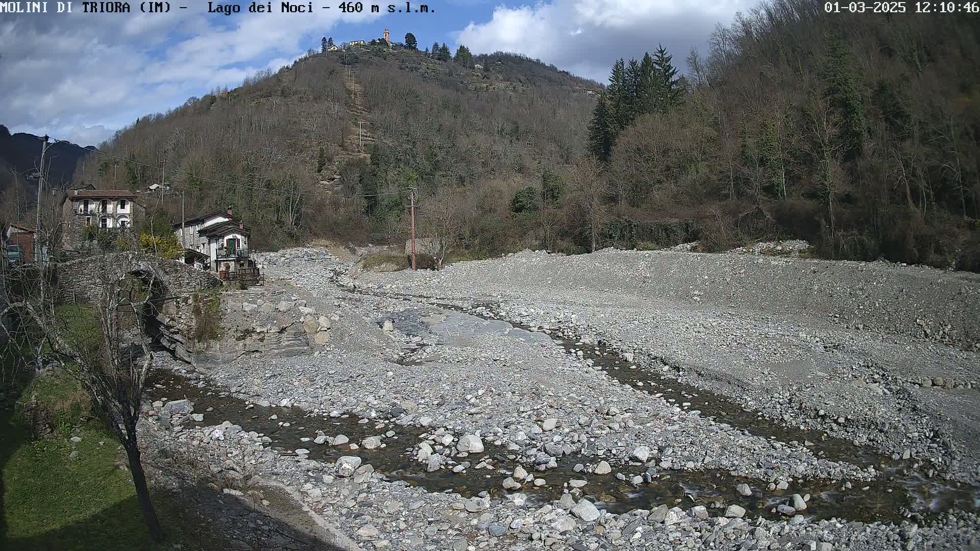 A rocky, mostly dry riverbed with a small stream flows through it, past a stone bridge and buildings nestled against a hillside under partly sunny skies.