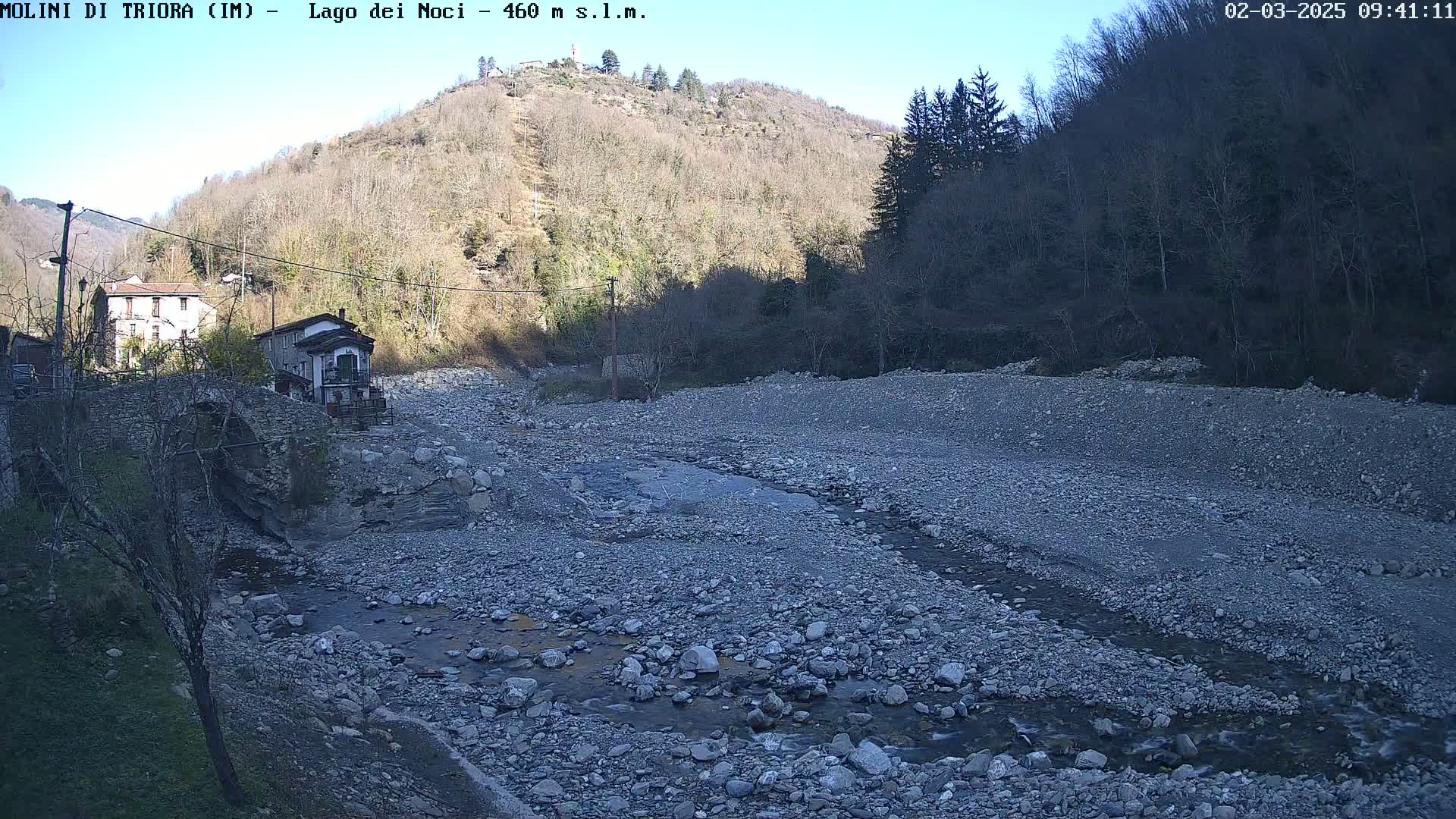 A rocky, dry riverbed with a small stream flows through it, next to buildings and a hill under a clear sky.