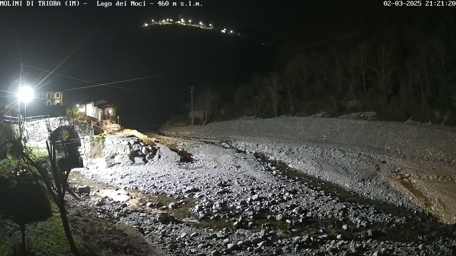 A nighttime view of a stone bridge over a shallow, rocky stream, illuminated by artificial lights, with a hill in the background.