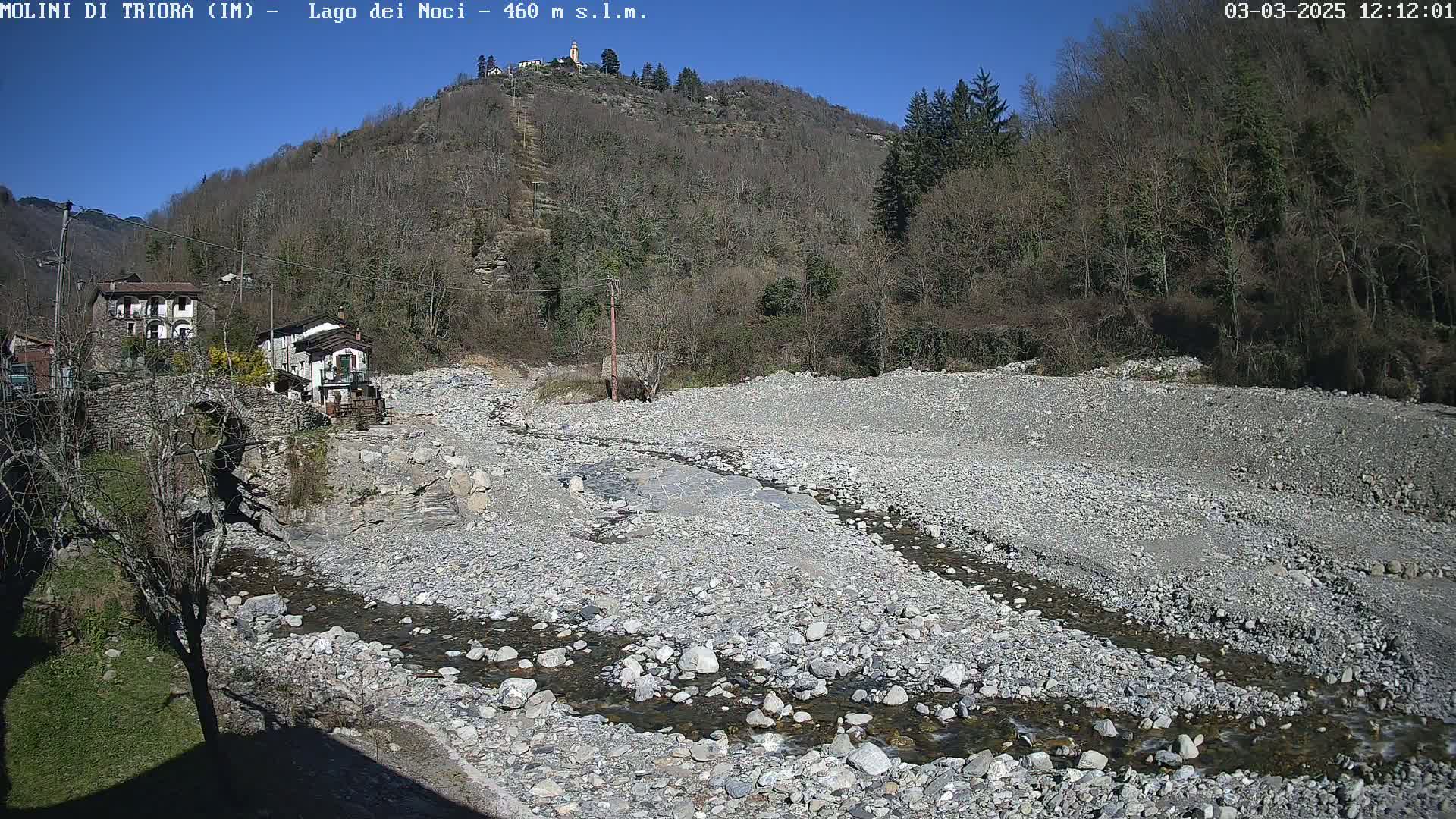 A rocky, mostly dry riverbed with a small stream flows through it, past a stone bridge and village nestled against a hillside under a clear, sunny sky.