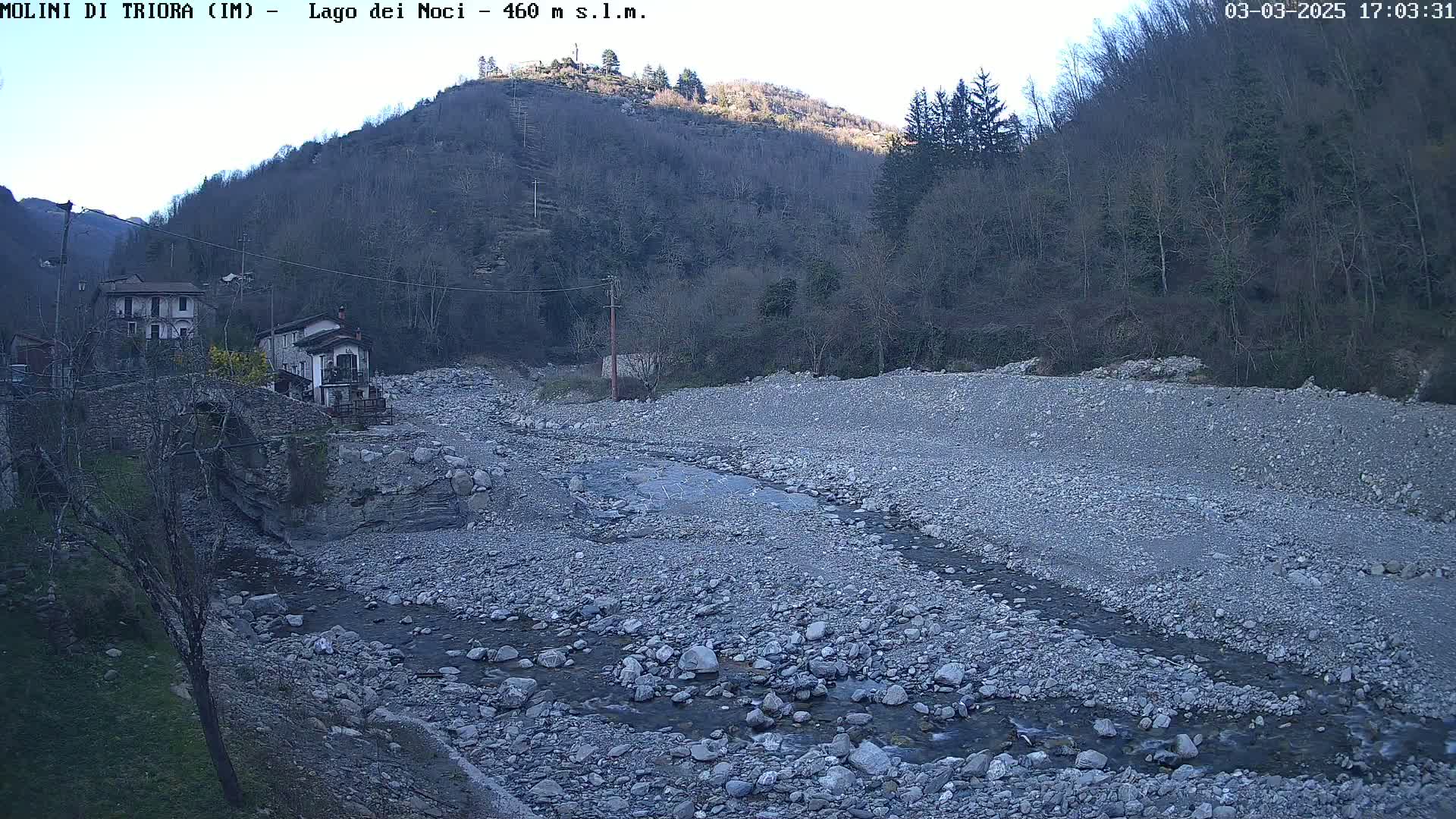 A rocky, dry riverbed with a small stream flows through it, past a stone bridge and houses nestled against a hillside under a clear, twilight sky.