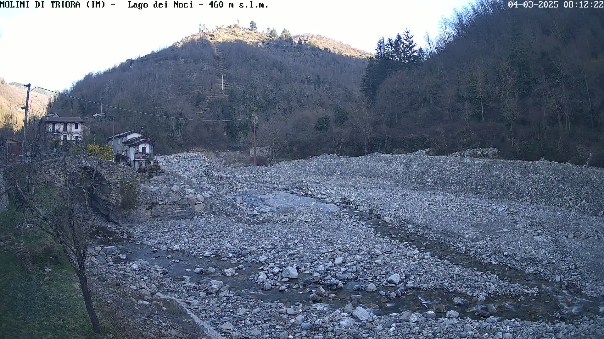 A rocky, dry riverbed with a small stream flows through it, near a village nestled against a hillside under a mostly clear sky.