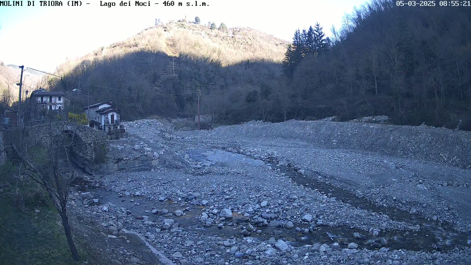 A rocky, dry riverbed with a small stream flows through it, past a stone bridge and houses nestled against a hillside under a clear, sunny sky.
