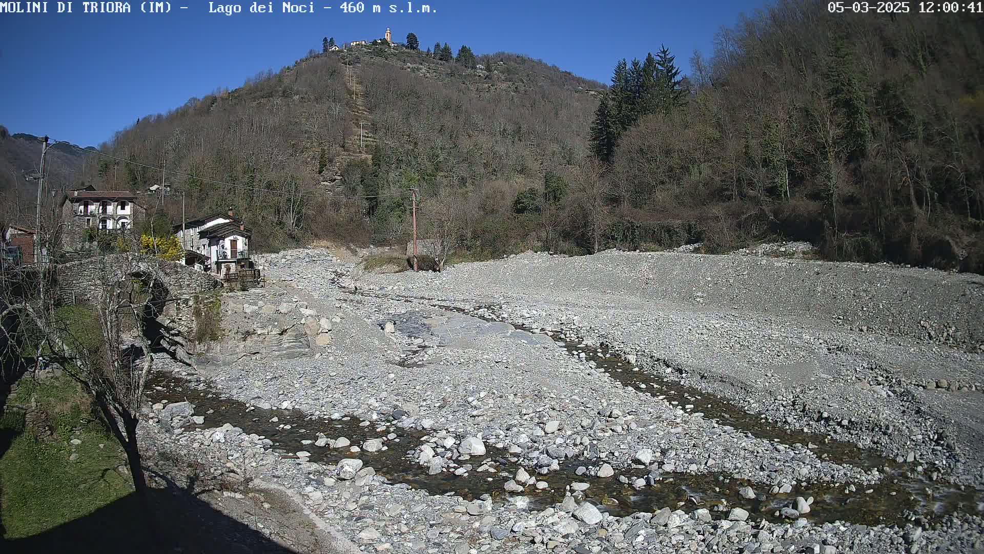 A rocky, dry riverbed with a small stream flows through it, nestled in a valley below leafless trees under a clear blue sky.