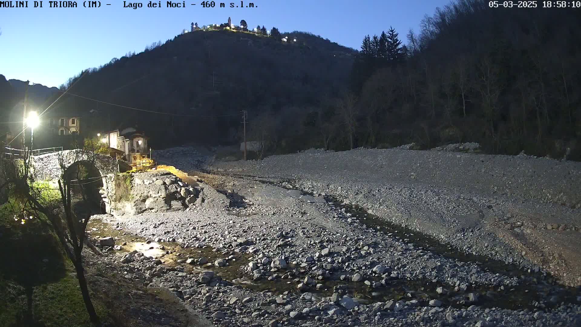 A rocky, dry riverbed with a small stream flows through it at dusk, under a hill with buildings and trees.