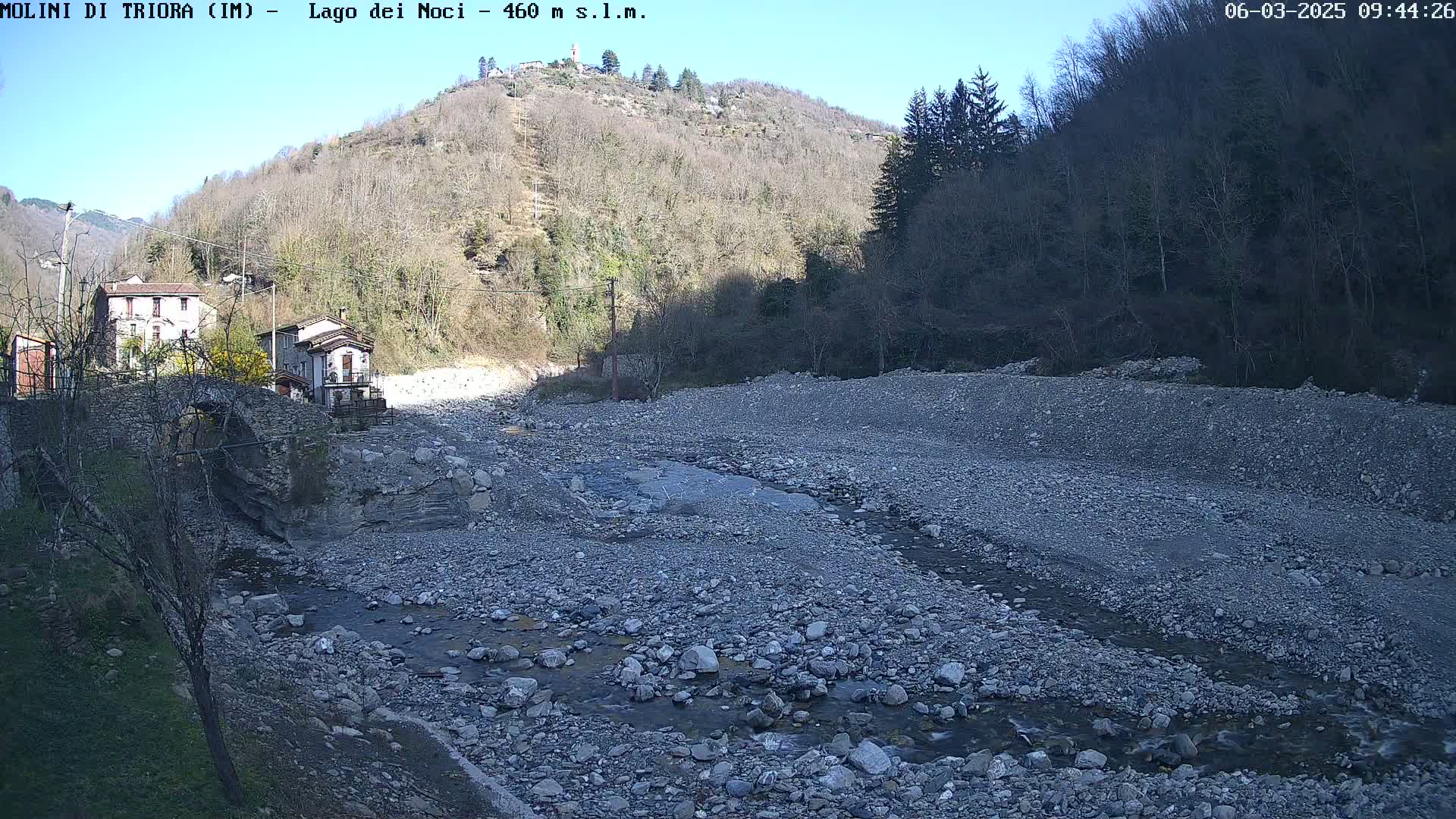 A rocky, dry riverbed with a small stream of water flows through it, past a stone bridge and buildings nestled at the base of a tree-covered hill under a clear, sunny sky.