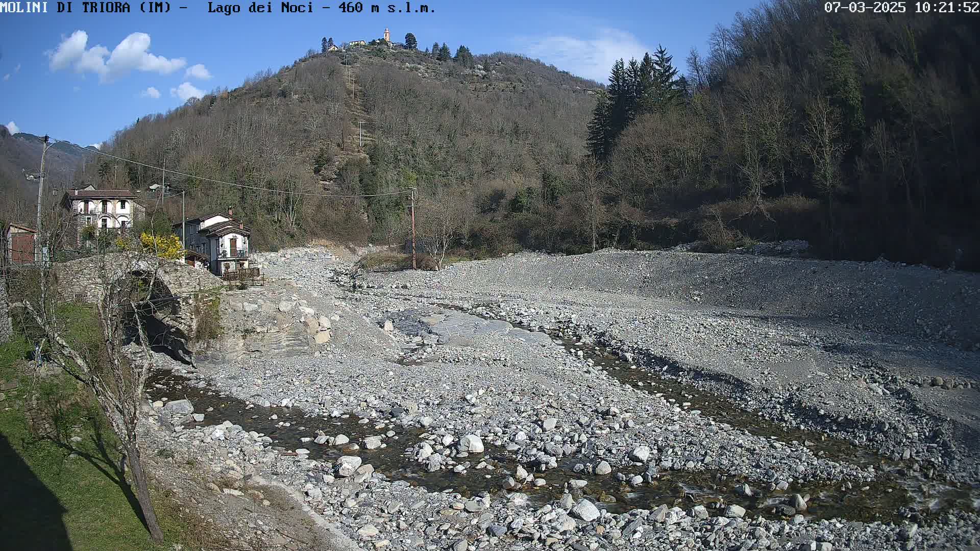 A mostly dry rocky riverbed with a small stream flows past a stone bridge and houses nestled against a hillside under a mostly sunny sky.
