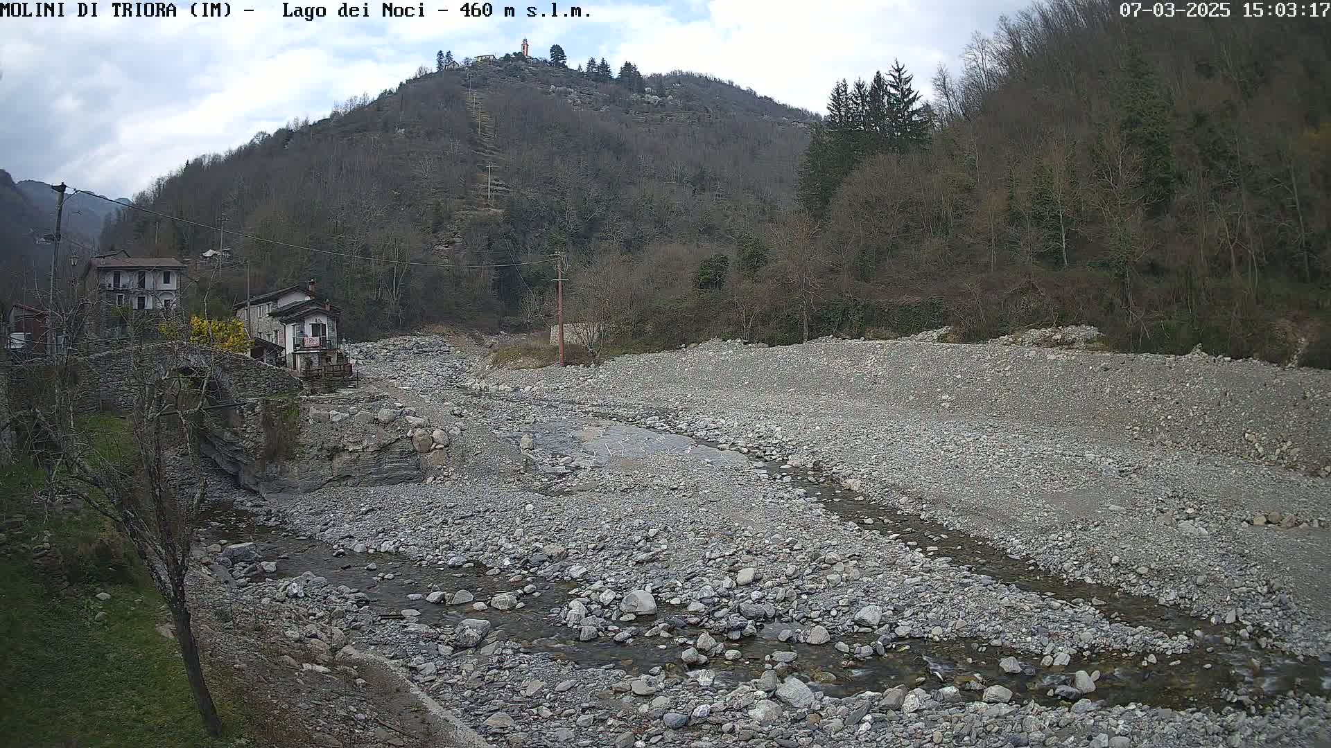 A rocky, mostly dry riverbed with a small stream flows through it, near a village nestled at the foot of a sparsely wooded hill under a partly cloudy sky.