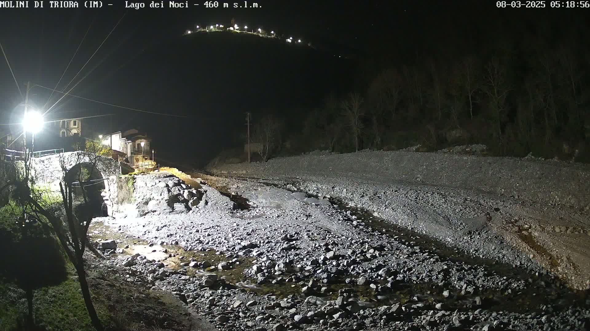 A nighttime view of a rocky stream bed with a stone bridge and buildings visible in the background under artificial light.