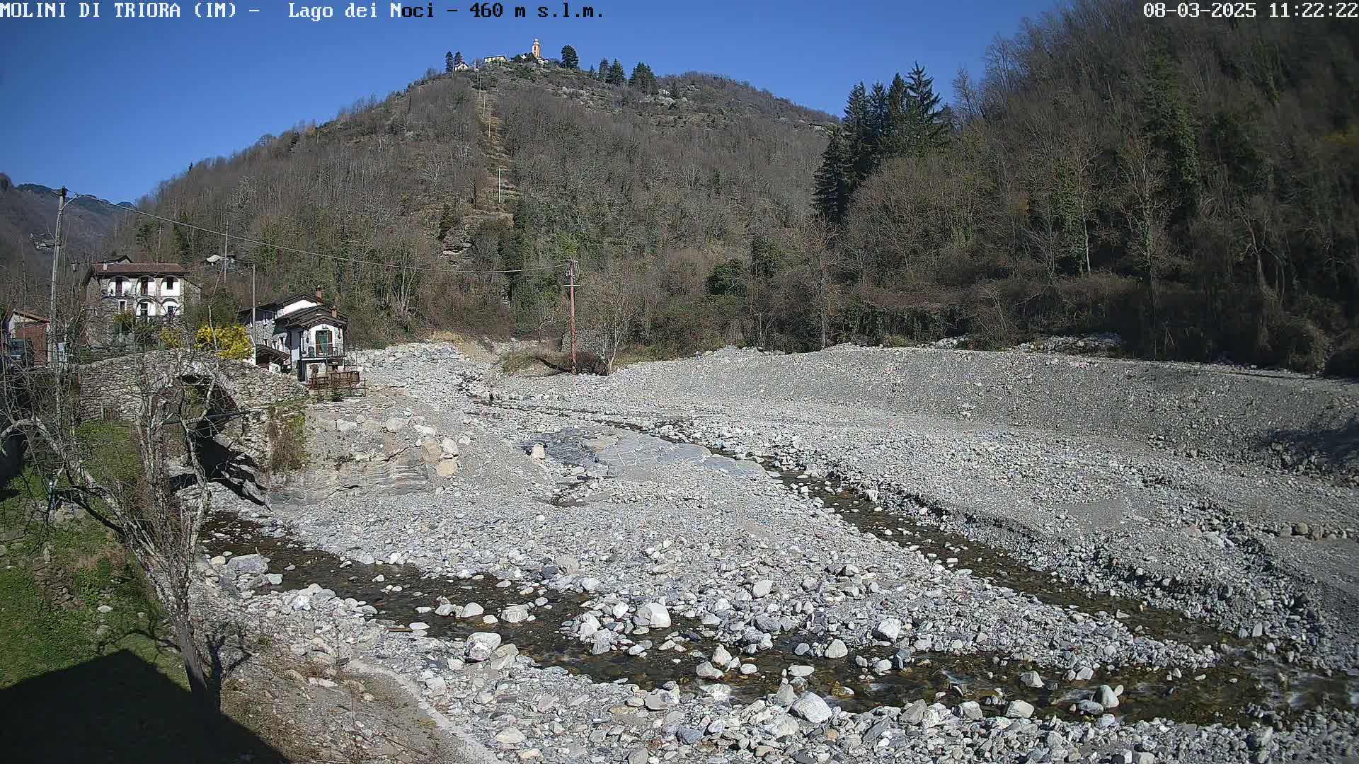 A rocky, mostly dry riverbed with a small stream flows through it, in front of a village nestled against a hillside under a clear blue sky.