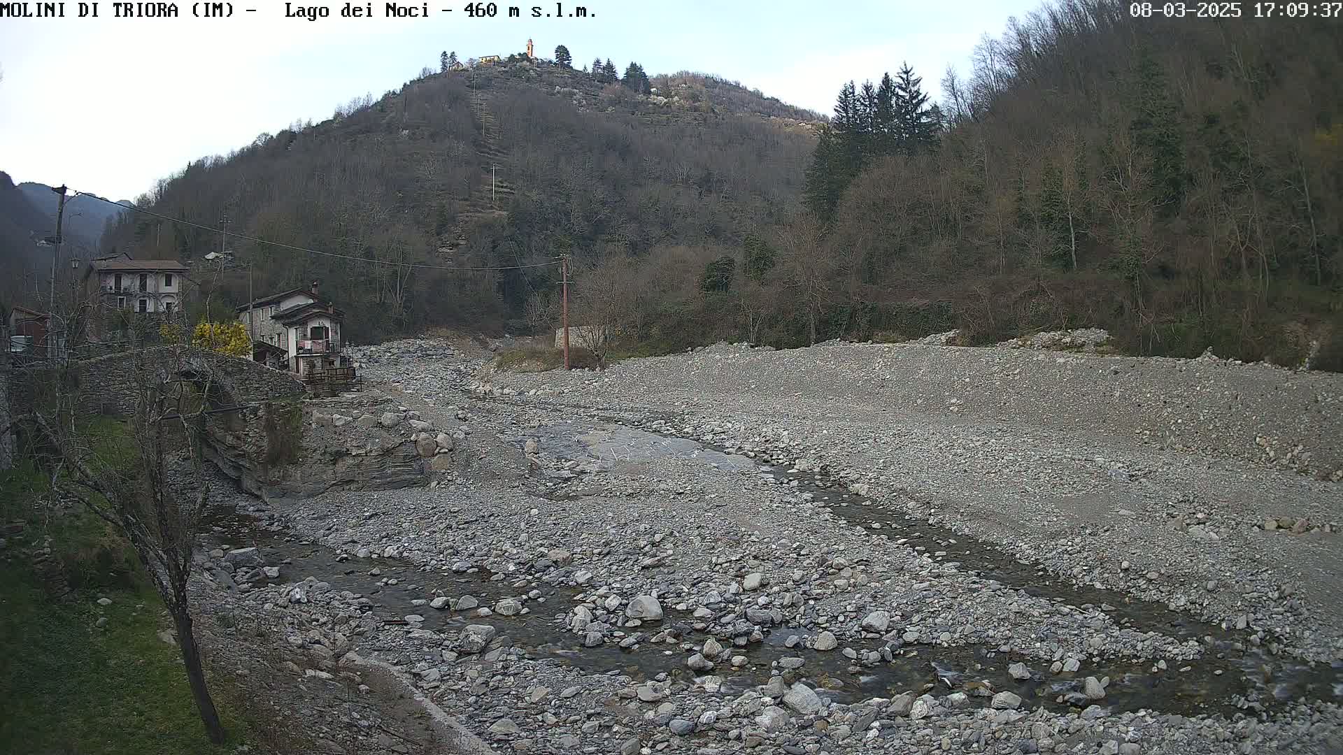 A rocky, dry riverbed with a small stream flows through it, next to a village nestled against a hillside under a mostly cloudy sky.