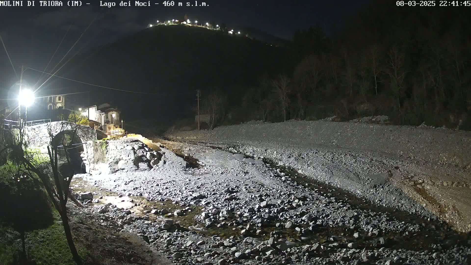 A rocky, shallow stream flows under a stone bridge at night, with a village and a hill visible in the background under a clear sky.