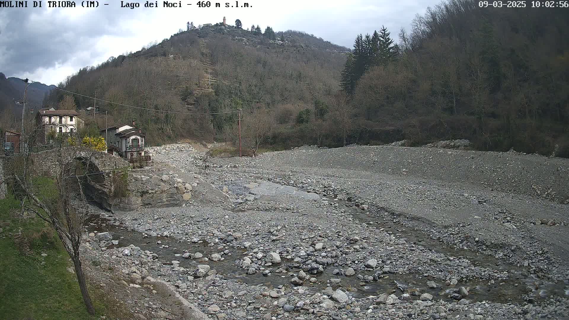 A rocky, mostly dry riverbed with a small stream flows past a stone bridge and several buildings nestled against a hillside under an overcast sky.