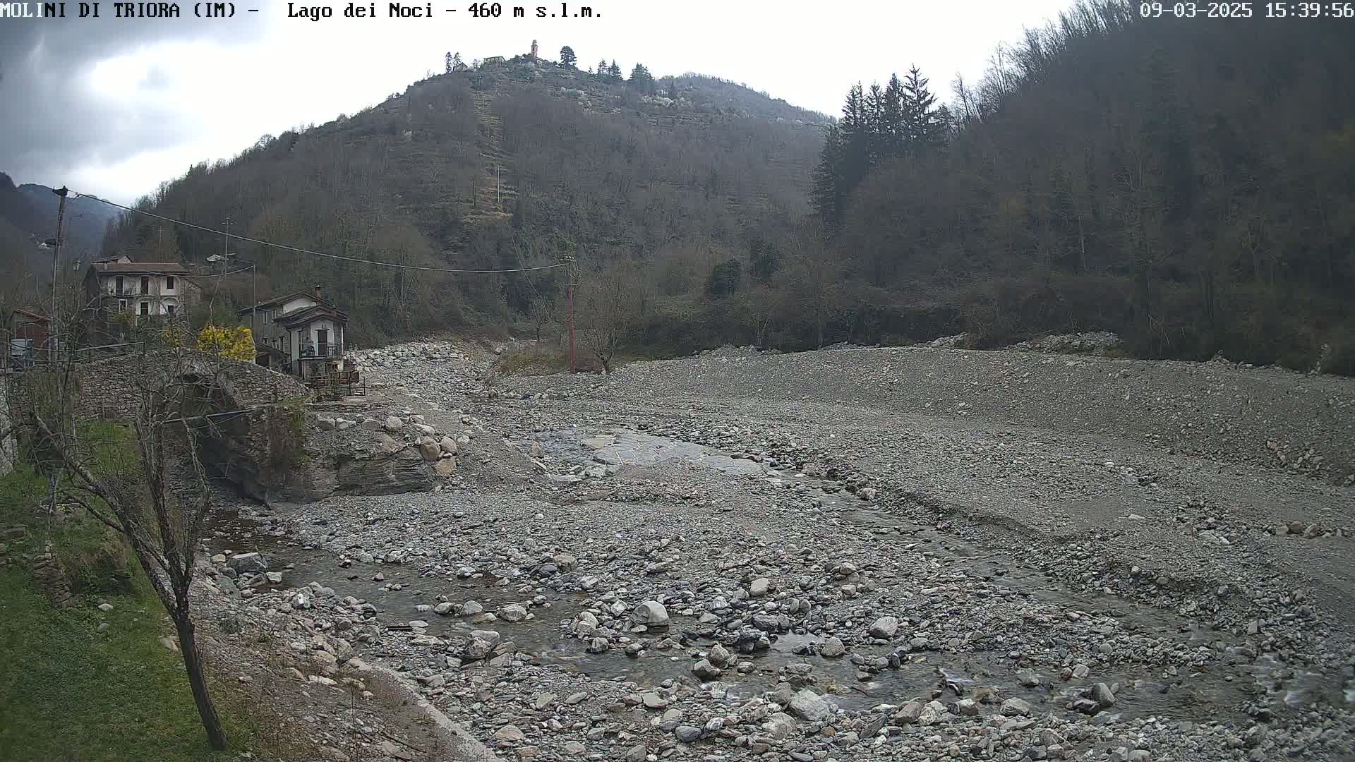 A rocky, dry riverbed with a small stream of water flows through it, situated in front of a village nestled at the base of a hill covered in bare trees under an overcast sky.