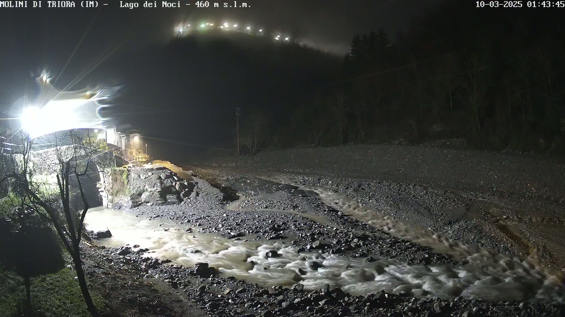 A nighttime scene shows a flooded river flowing rapidly over a rocky riverbed, partially undermining a stone bridge under foggy conditions.