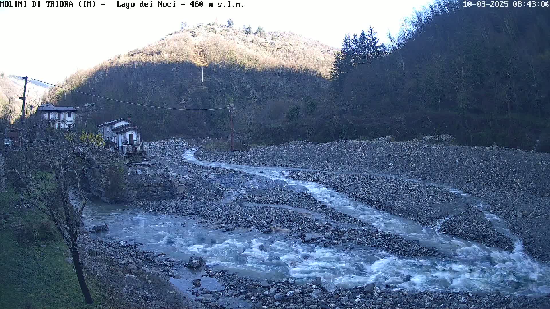 A rocky river flows through a valley, past a few houses nestled at the base of a hill under a clear, sunny sky.