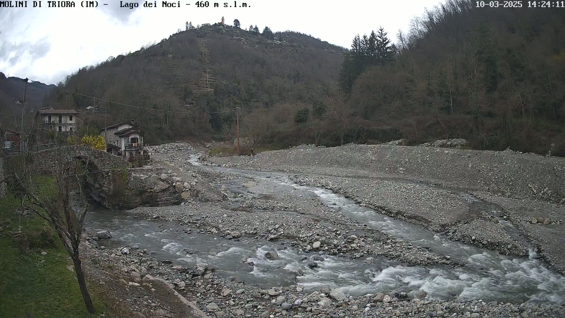 A rocky river flows through a valley under an overcast sky, past a stone bridge and small village nestled against a hillside.
