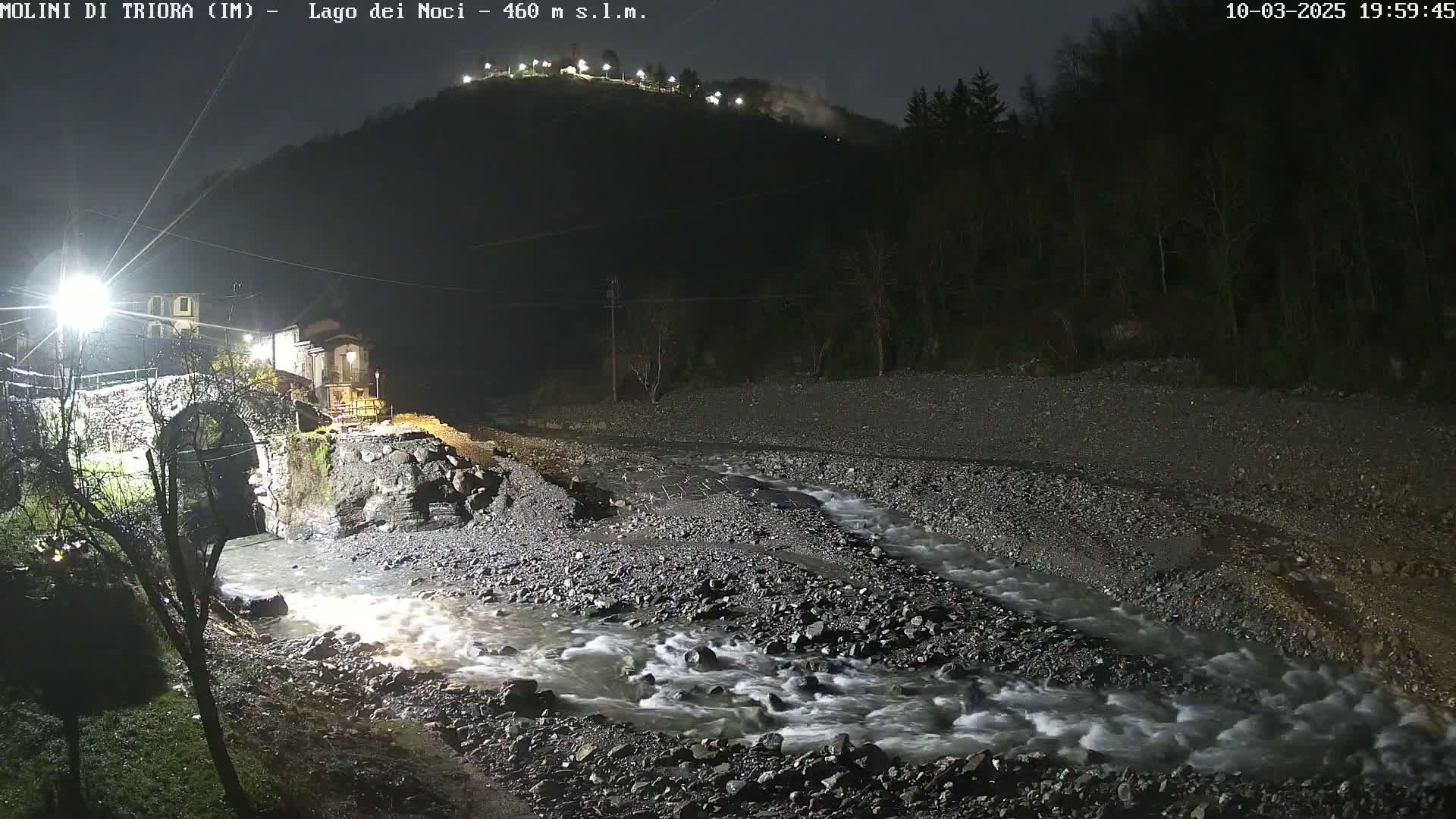 A nighttime, floodlit view of a rocky river flowing past a stone bridge and small village nestled at the base of a hill under a dark sky.