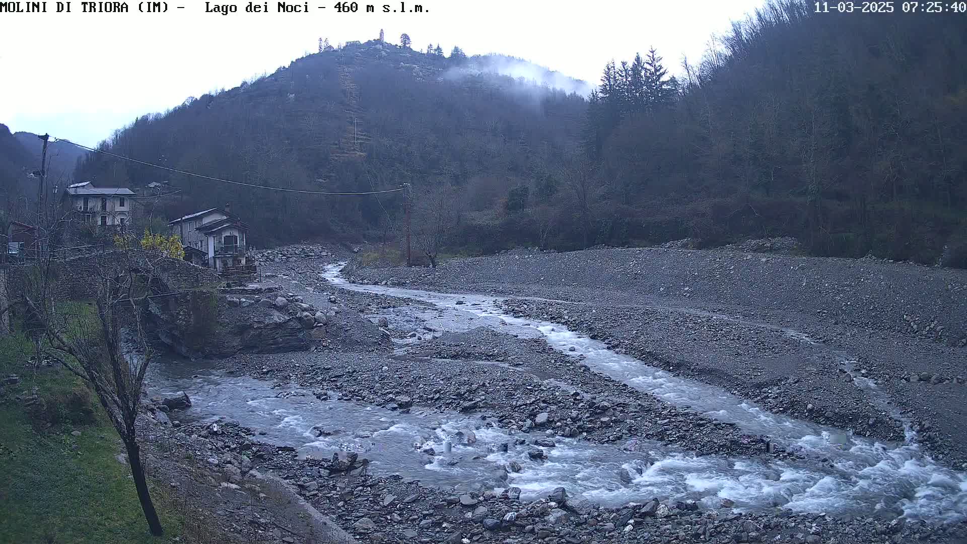 A rocky river flows through a valley, past a small village nestled against a misty hillside under an overcast sky.