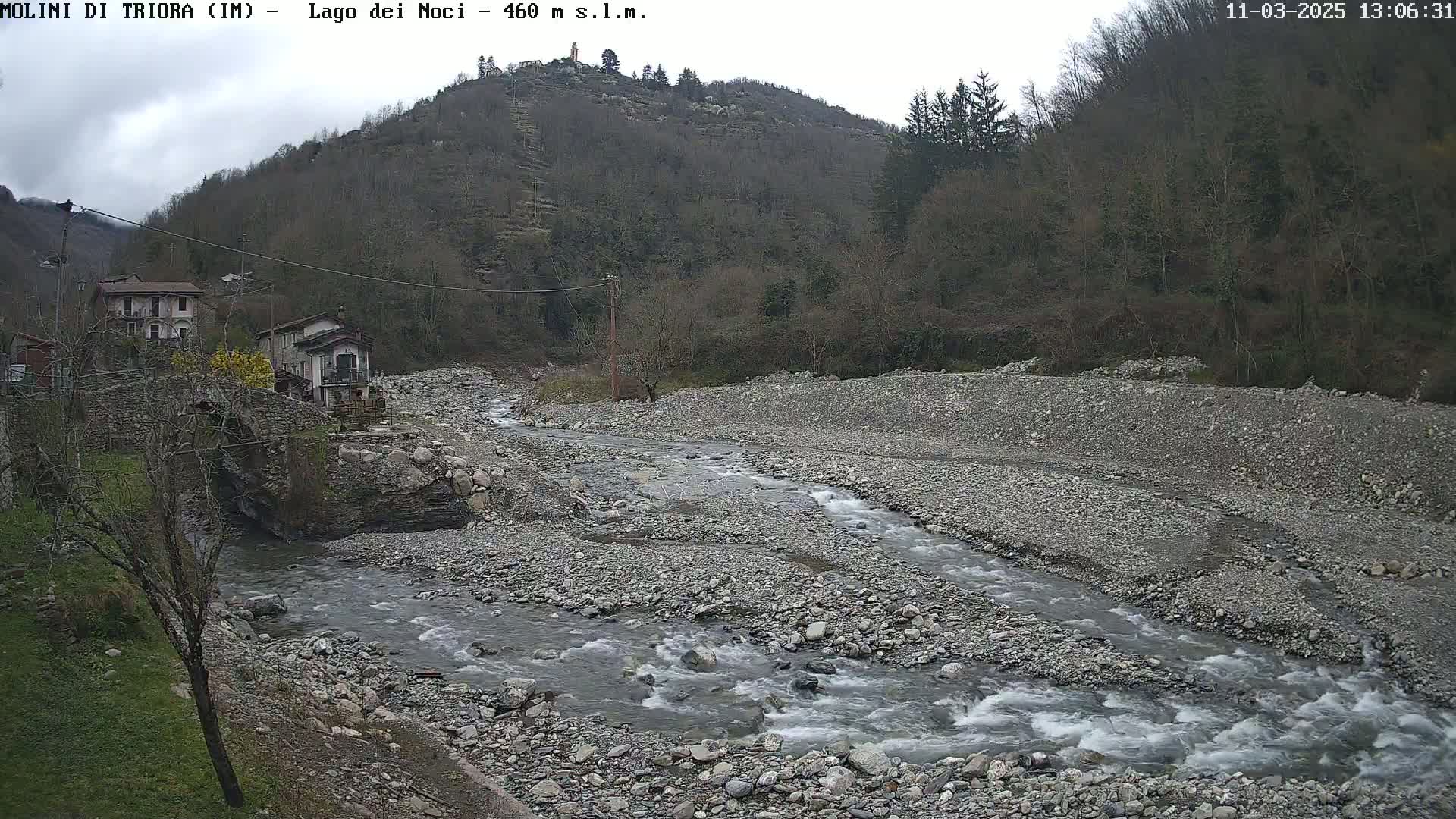 A rocky river flows through a sparsely vegetated valley under an overcast sky, past a stone bridge and small buildings nestled against a hillside.