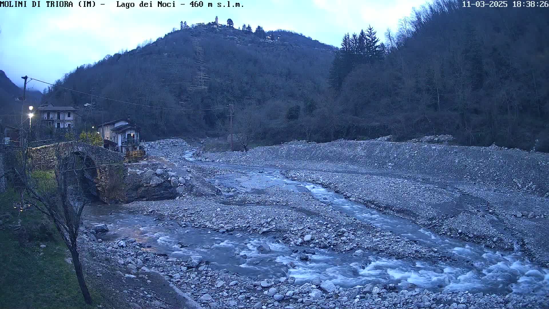 A rocky river flows past a stone bridge and small village nestled in a valley under a twilight sky.