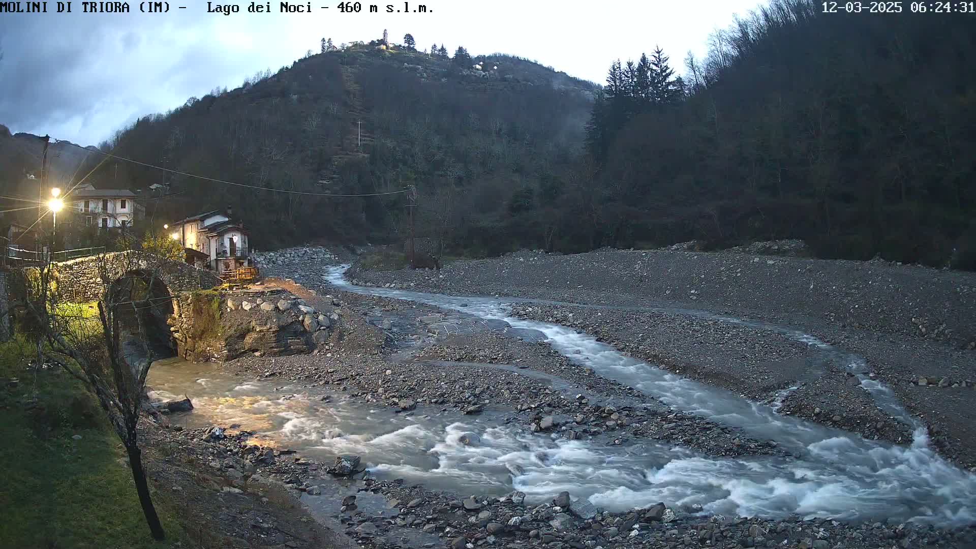 A rocky river flows under a stone bridge near a small village nestled in a valley, under a twilight sky.