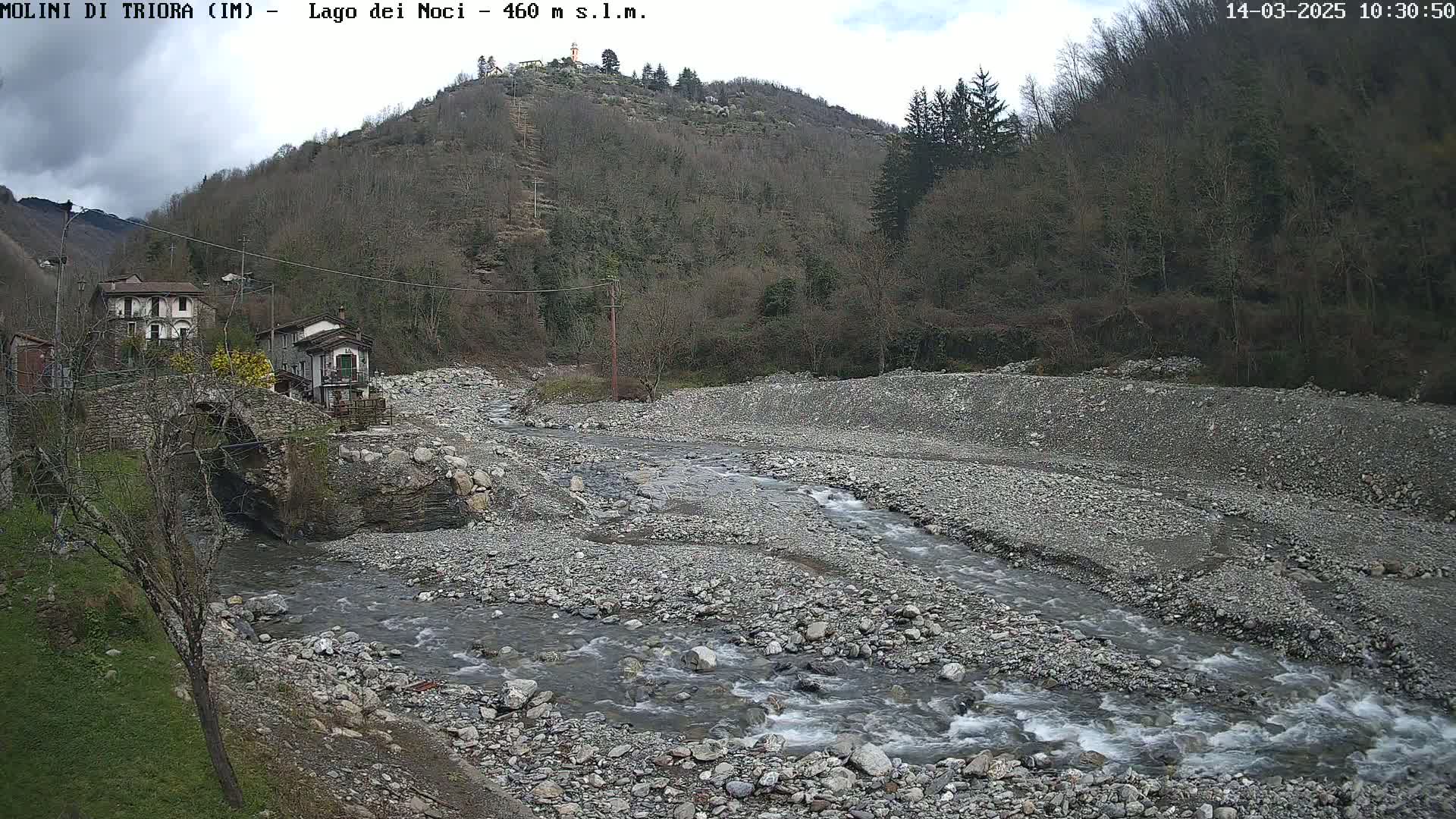 A rocky river flows through a valley under a partly cloudy sky, past a stone bridge and a small village nestled against a hillside covered in leafless trees.