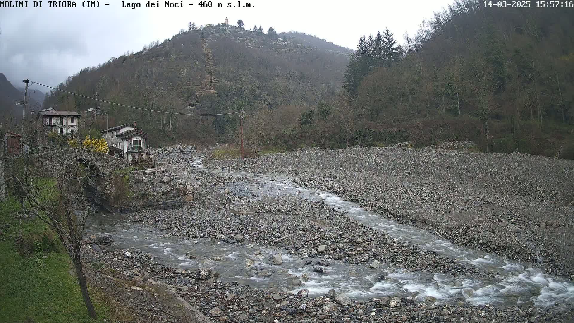 A rocky, gray river flows through a sparsely vegetated valley under an overcast sky, past a small village with stone buildings and a stone bridge.