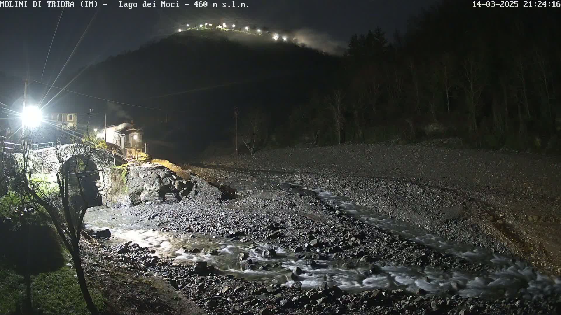 A rocky stream flows under a stone bridge at night, past a small village nestled at the base of a hill with lights visible on its upper slopes.