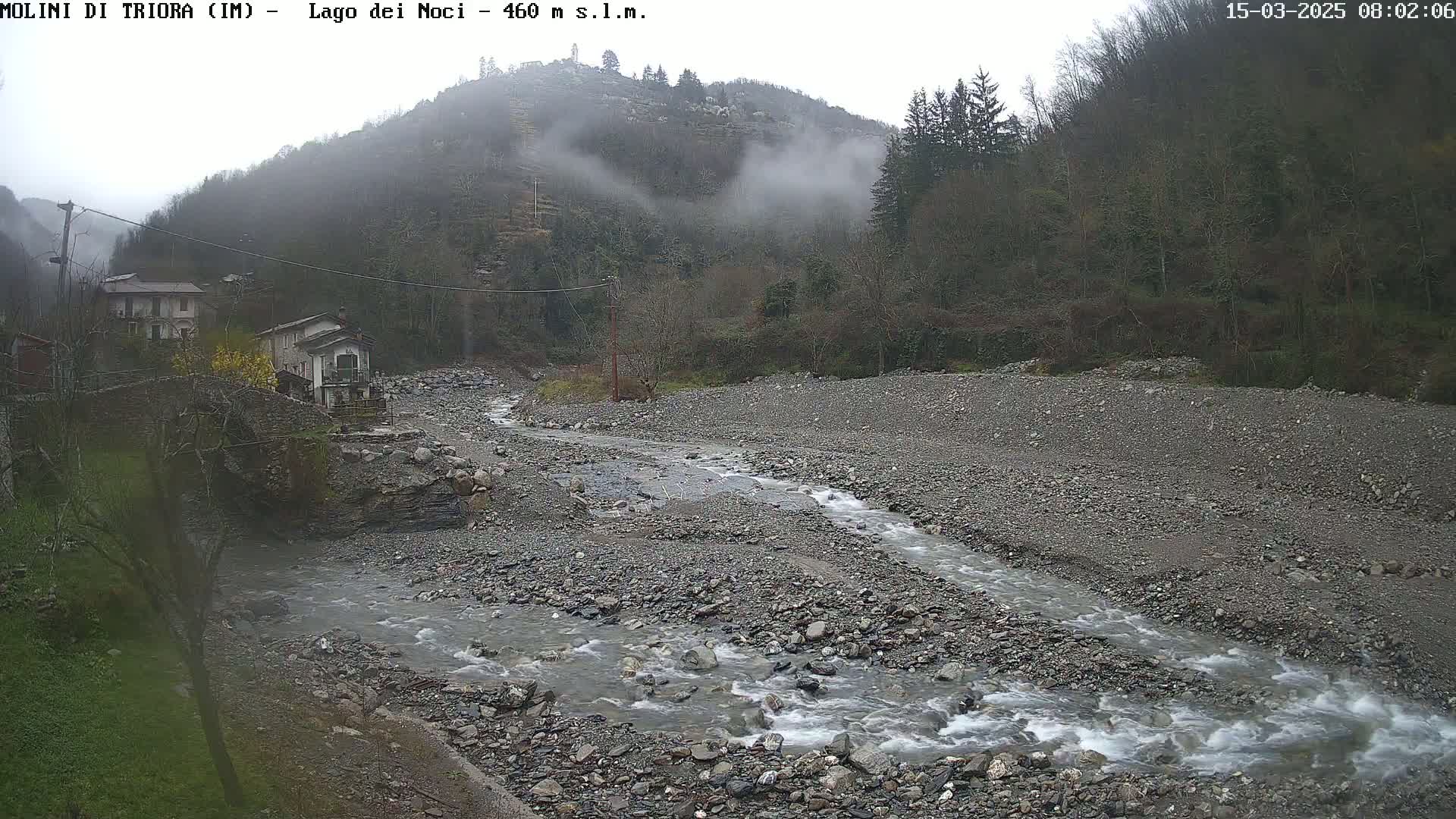 A rocky river flows through a valley shrouded in fog, past a small village nestled against a hillside.