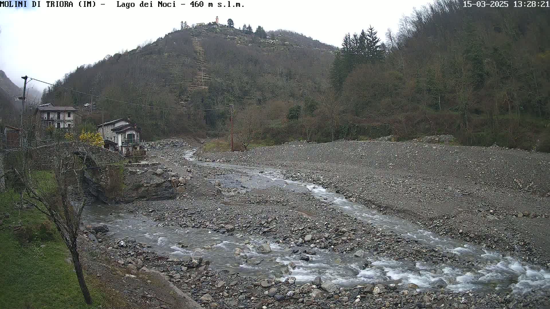 A rocky river flows through a valley, past a small village nestled at the base of a hill under an overcast sky.