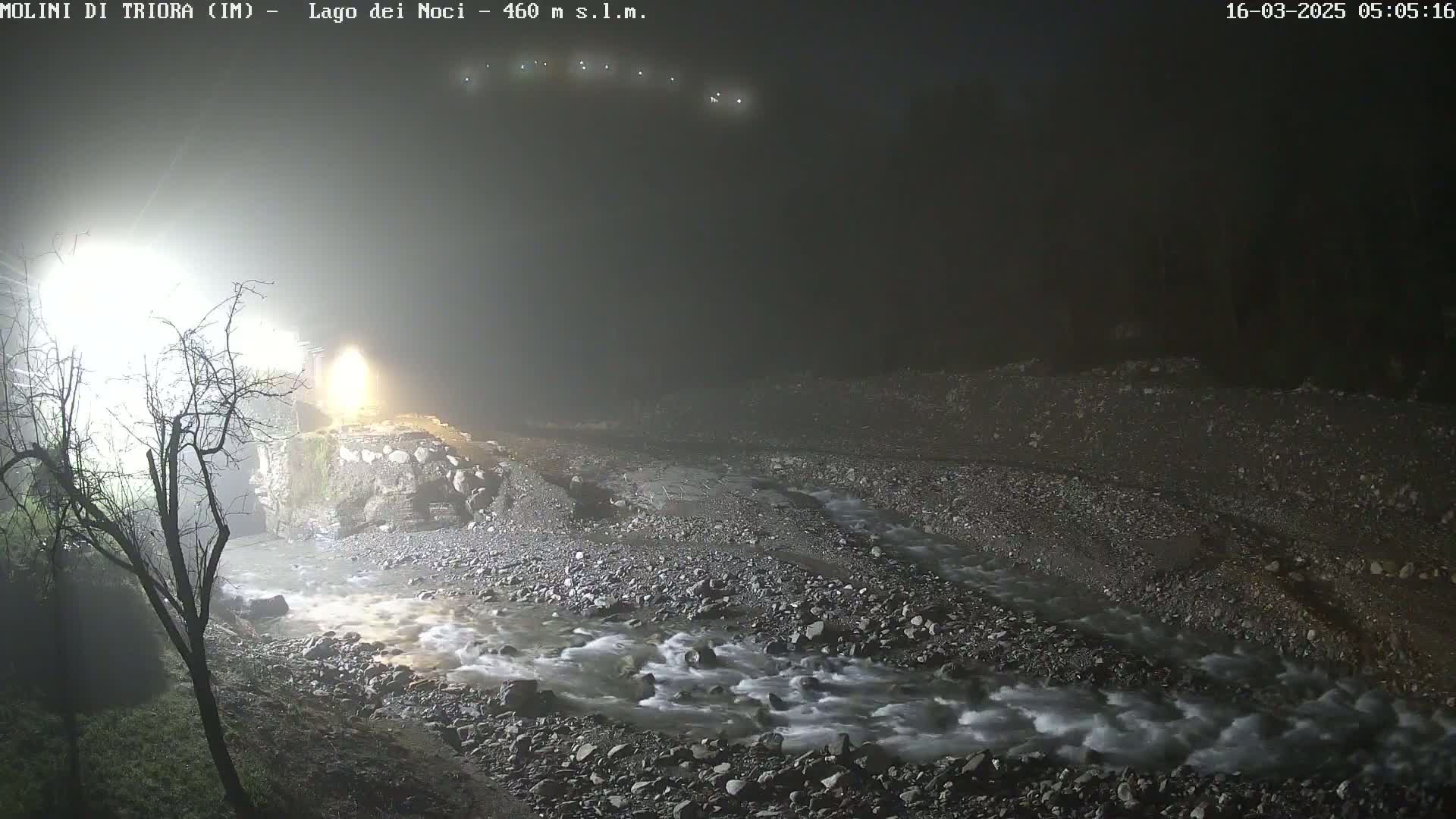 A rocky stream flows under a bridge at night in foggy conditions, illuminated by bright lights.