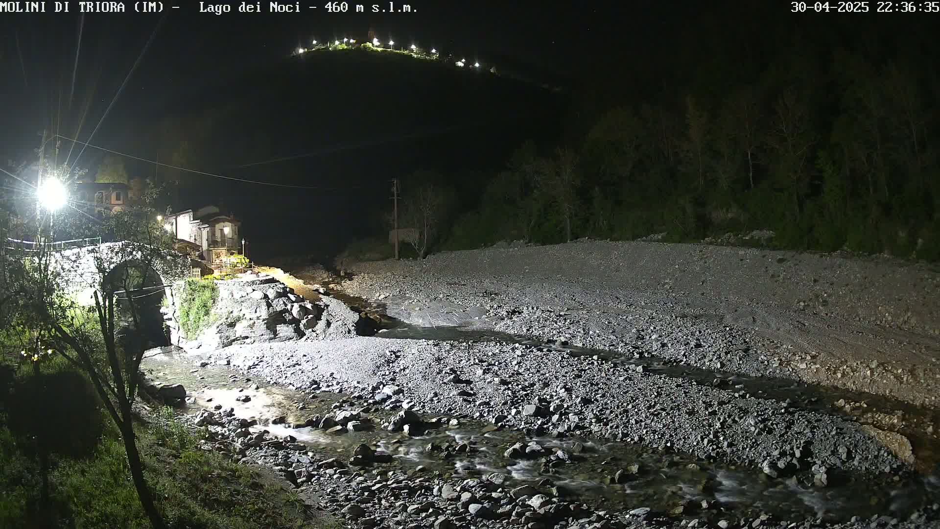 A rocky, shallow stream flows through a nighttime landscape, illuminated by artificial lights, past a stone bridge and buildings nestled against a hill.
