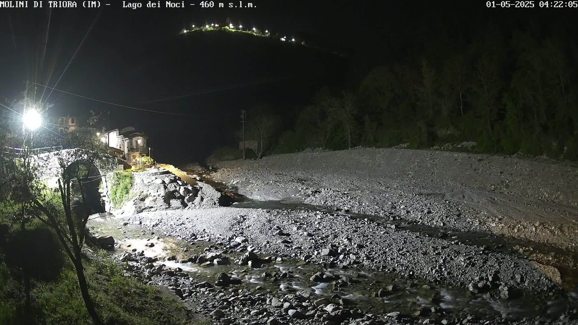 A rocky stream flows through a dry riverbed at night, past a stone bridge and a building, under a dark sky with lights illuminating the hillside in the distance.