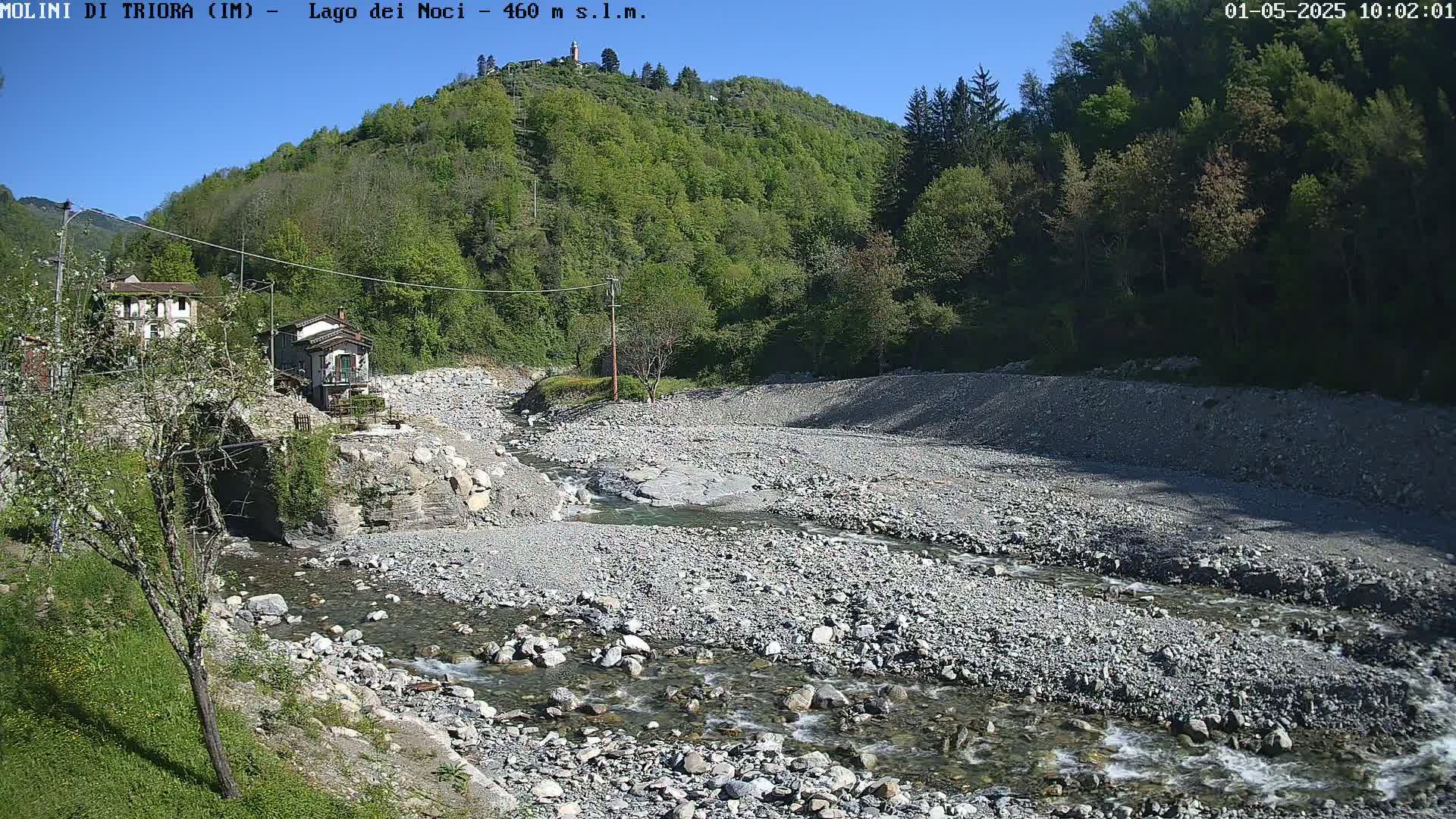 A rocky riverbed with a shallow, flowing stream winds through a valley under a clear, sunny sky, past small buildings and lush green hillsides.