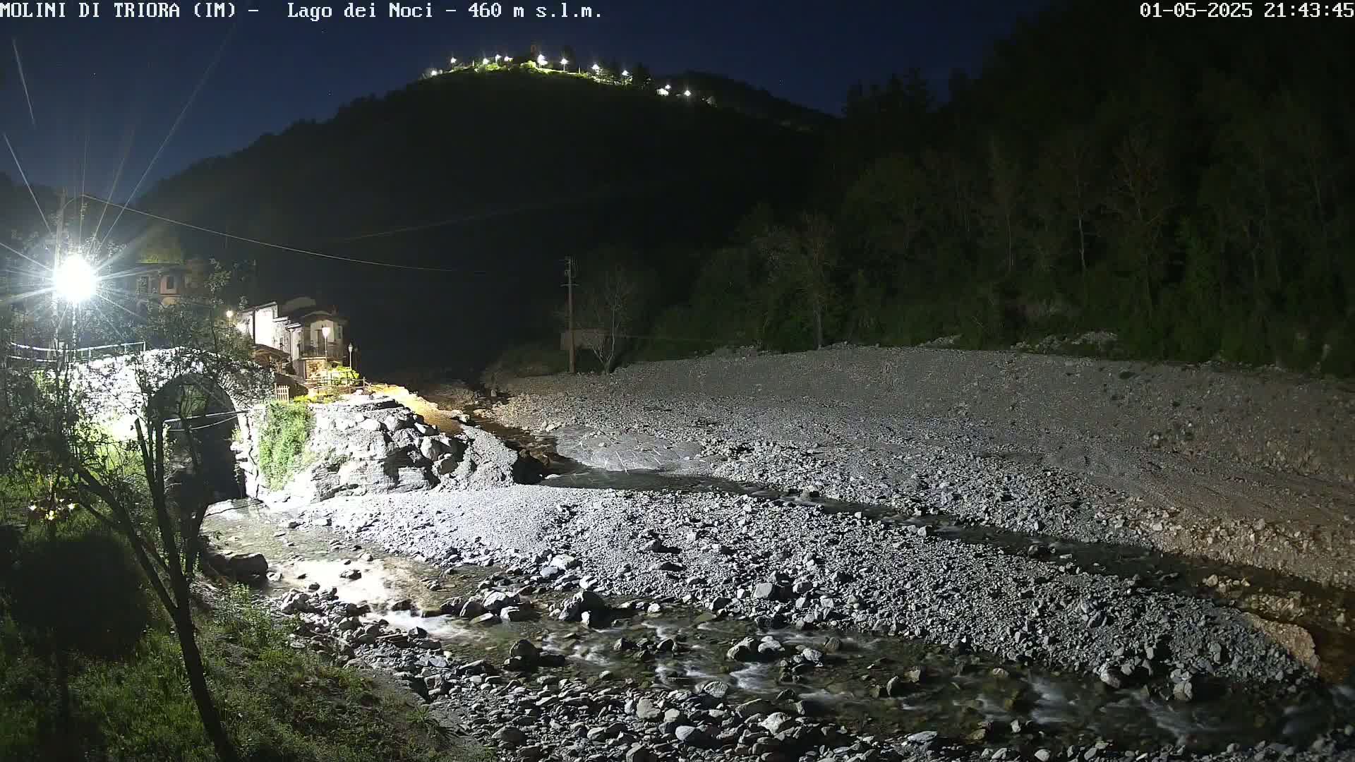 A rocky stream flows through a dimly lit valley at night, past a stone bridge and small village nestled beneath a hilltop with lights.