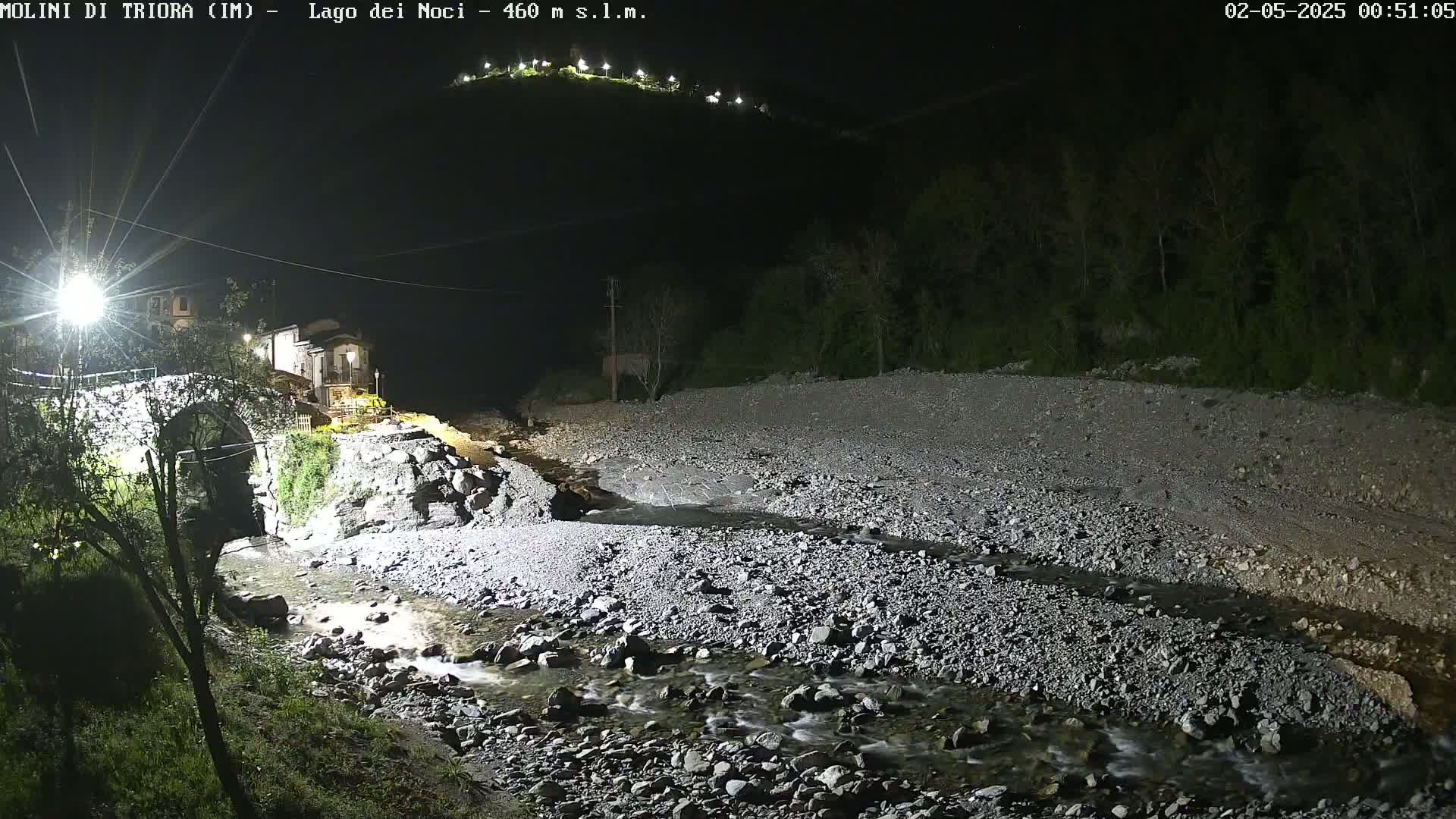 A rocky stream flows under a stone bridge at night, past a small village nestled at the base of a hill illuminated by lights.