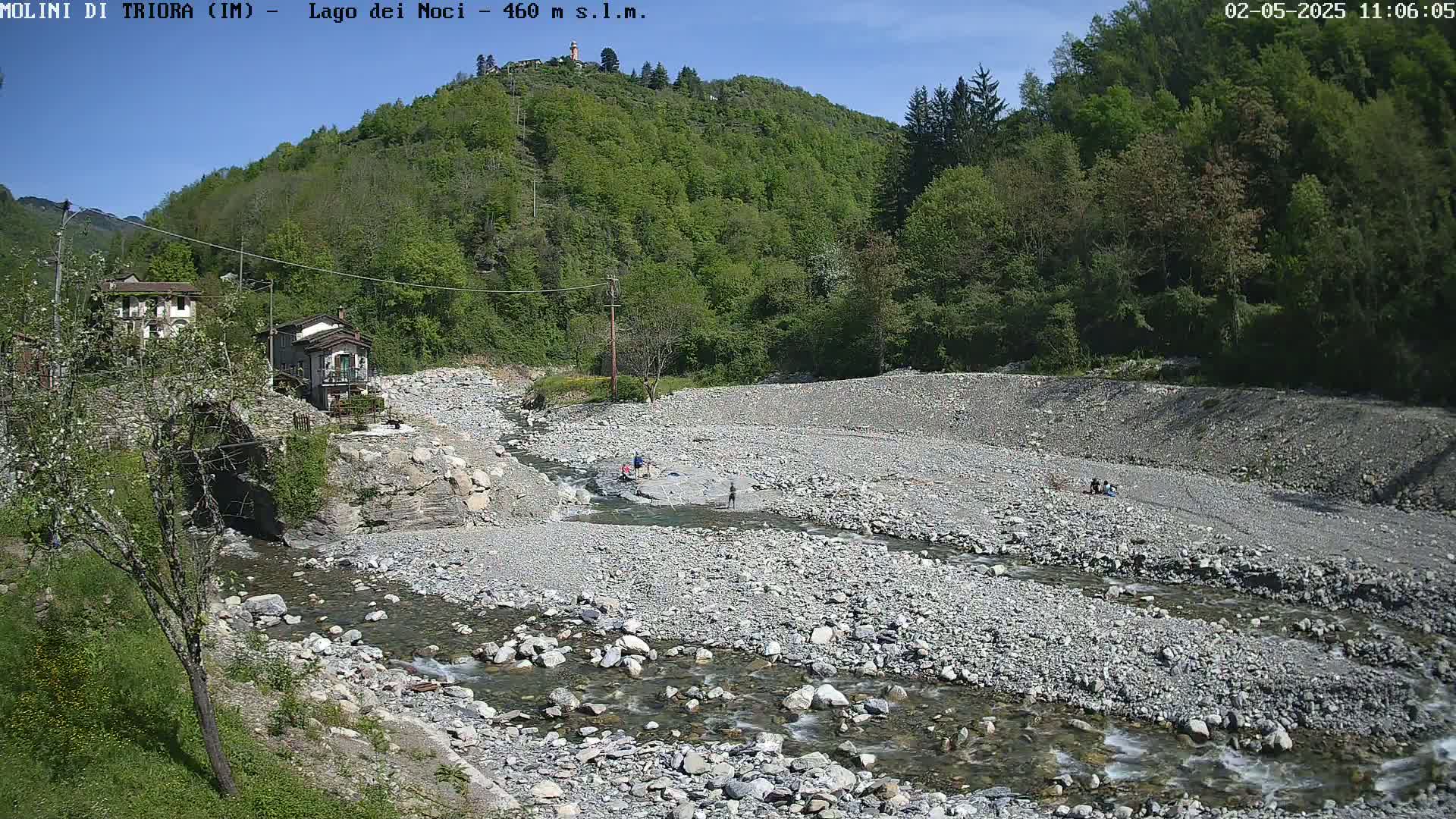 A rocky riverbed with a shallow, flowing stream winds through a valley under a sunny sky, past a few small buildings and people near the water's edge, surrounded by lush green hillsides.