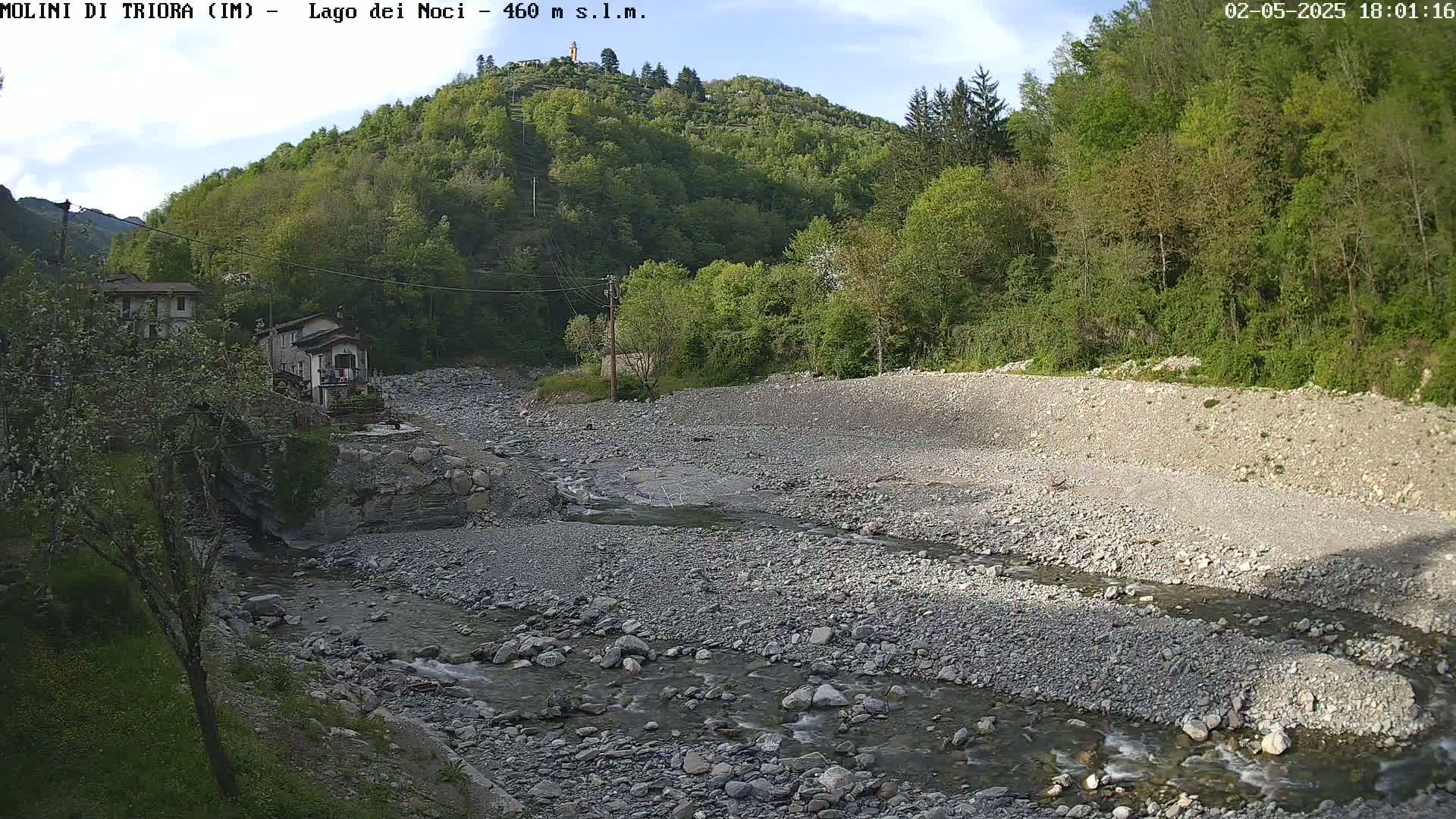 A rocky, shallow river flows through a dry, gravelly riverbed, surrounded by lush green trees and two small buildings under a partly cloudy sky.
