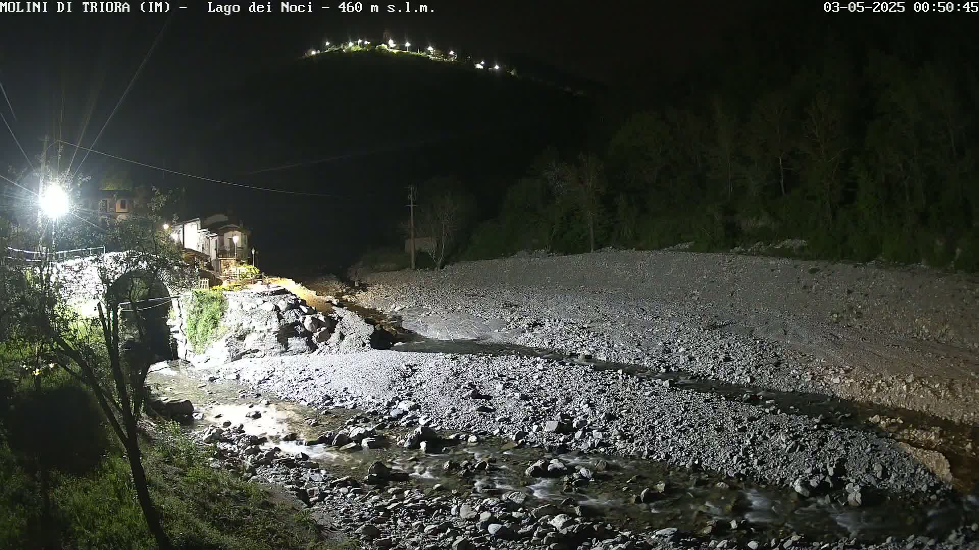 A rocky stream flows at night past a stone bridge and a small village nestled beneath a hill.