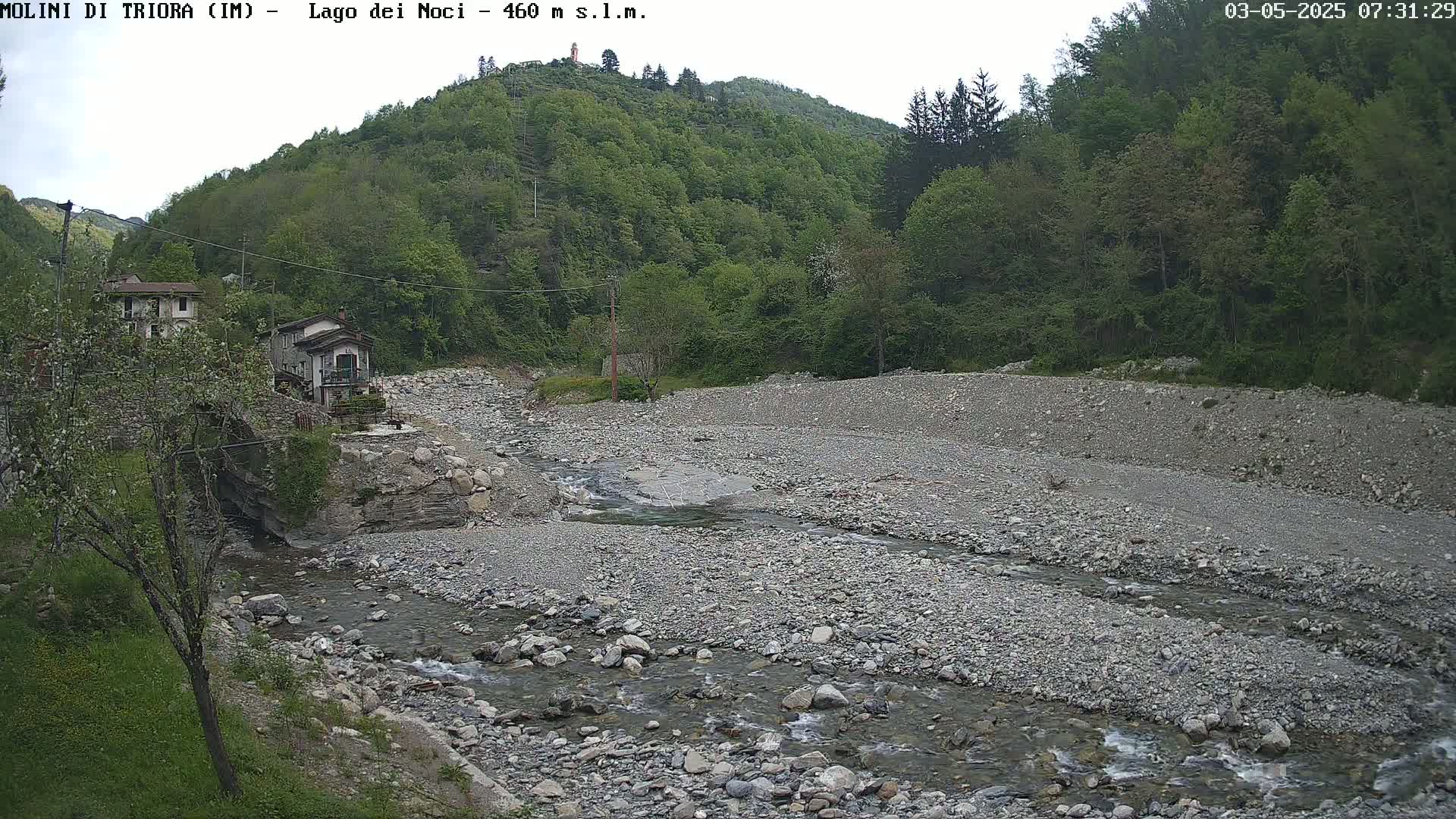 A rocky, mostly dry riverbed with a small stream flows through it, past buildings nestled against a green hillside under an overcast sky.
